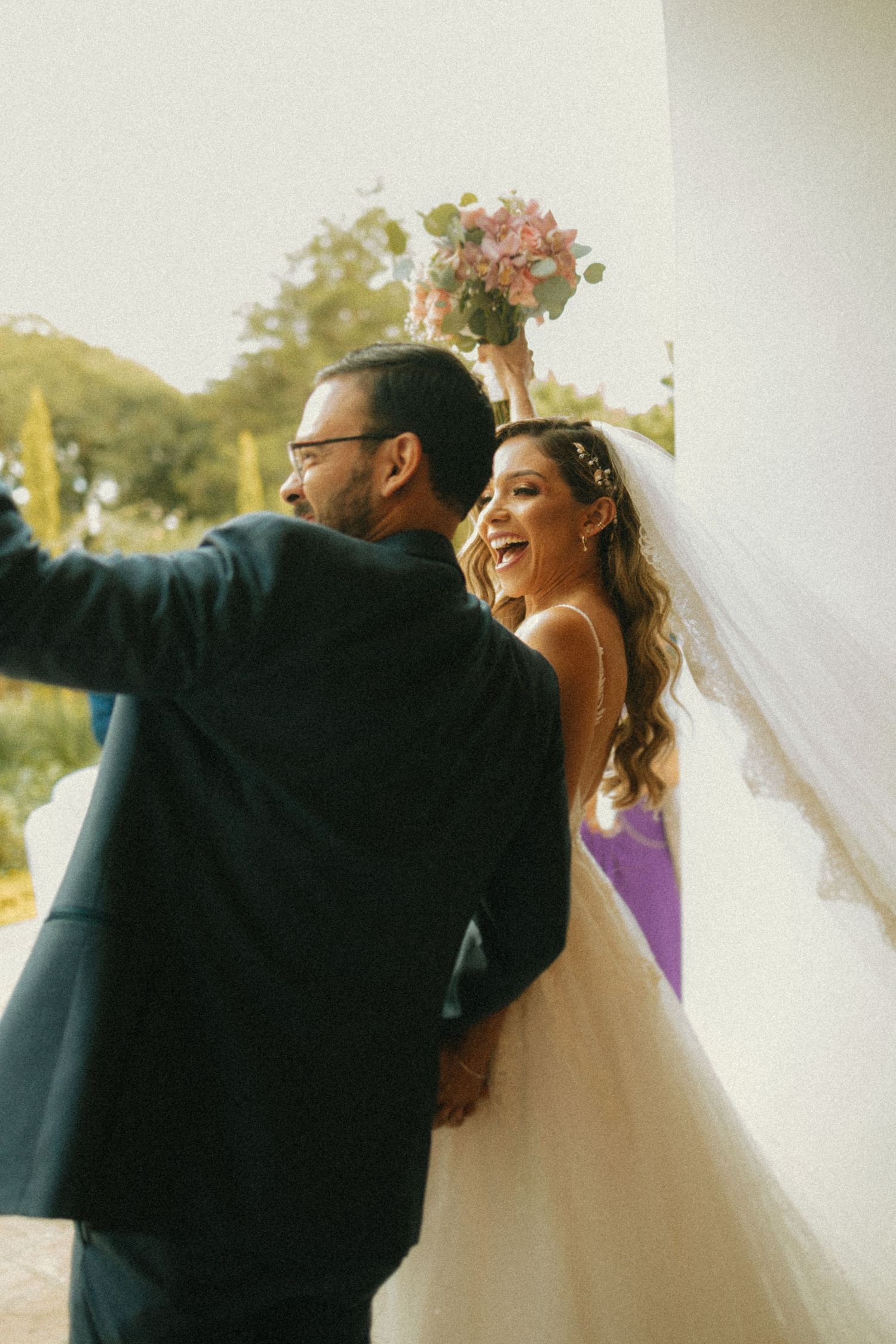 Joyful bride in white gown holding pink bouquet aloft laughs with groom in dark suit at outdoor wedding venue.
