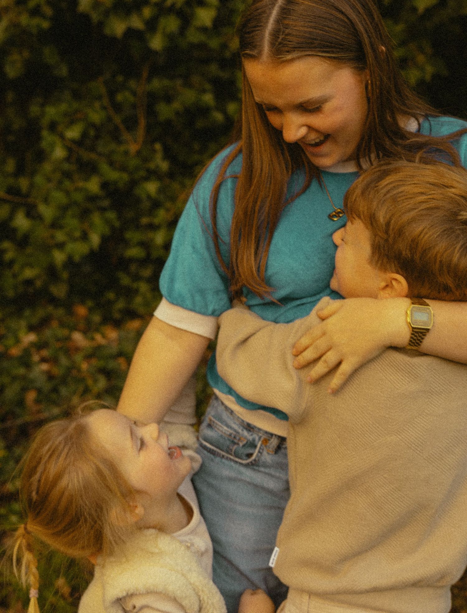 A teenage girl hugs two younger children outdoors, all smiling and laughing together.