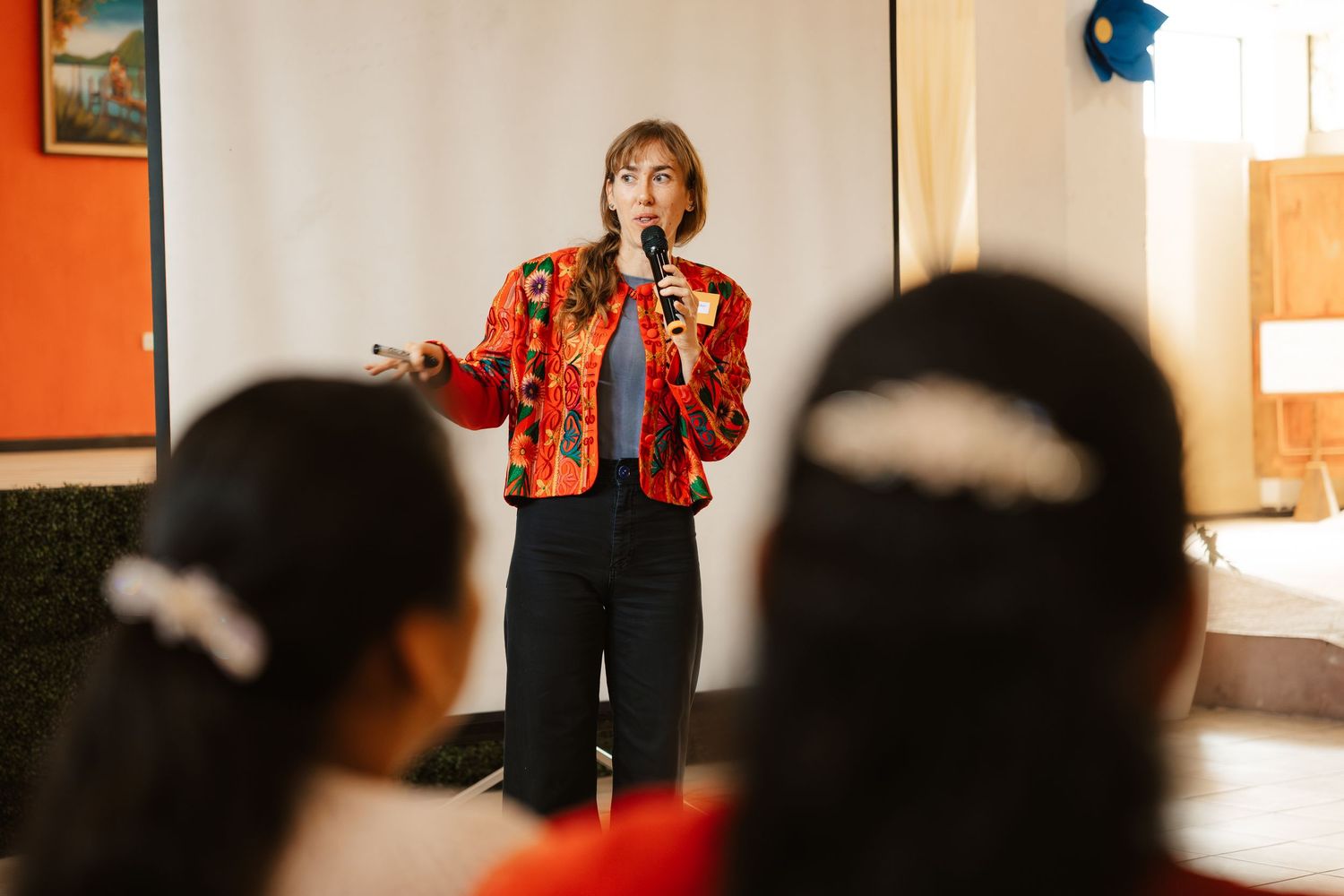 Woman in red floral jacket speaks into microphone while presenting to an audience at an indoor event.