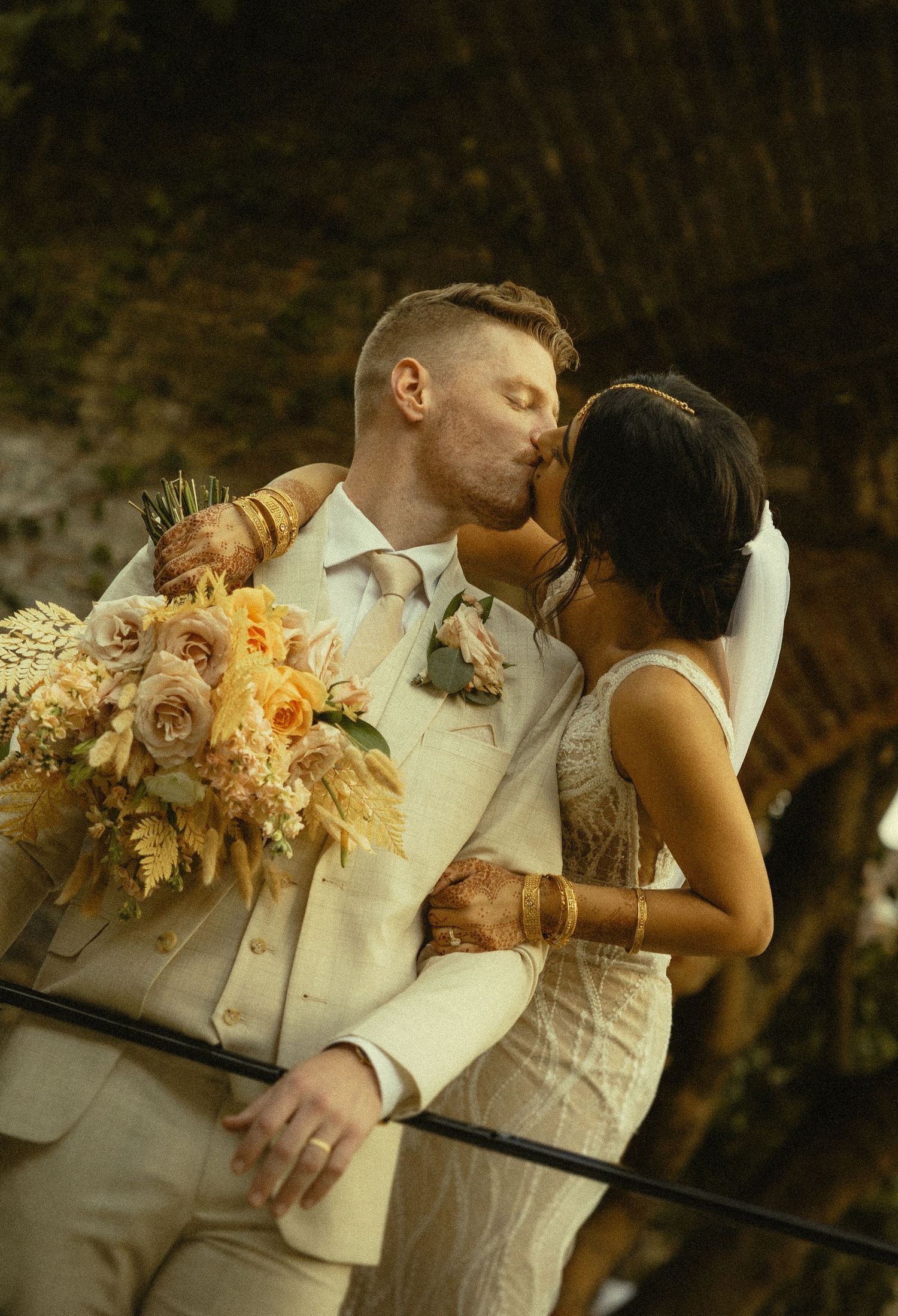 A couple shares a romantic kiss at their wedding, bride holding yellow floral bouquet with henna-adorned arms in elegant attire.