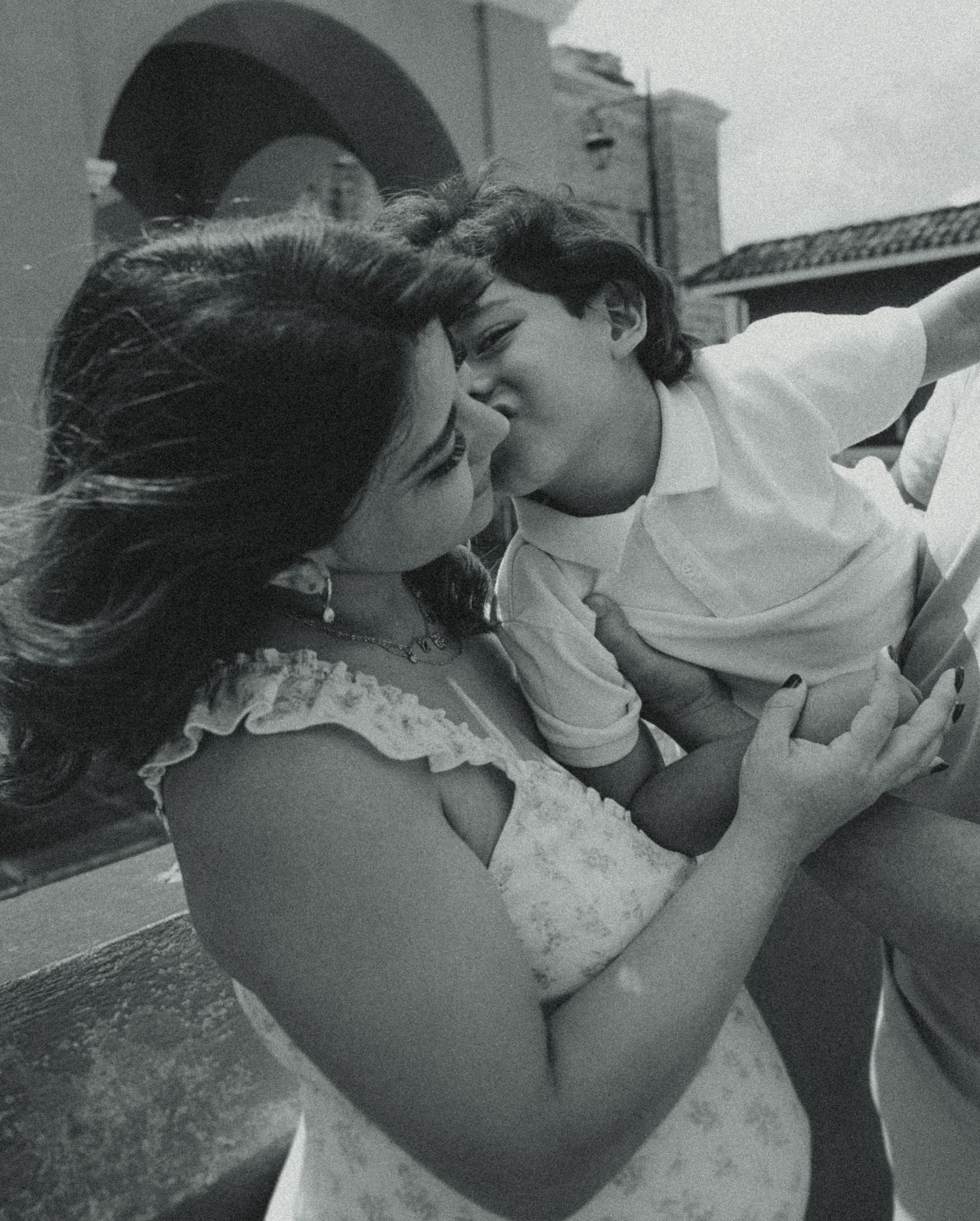 Black and white photo of a woman and young man sharing a joyful, affectionate moment outdoors near a building.