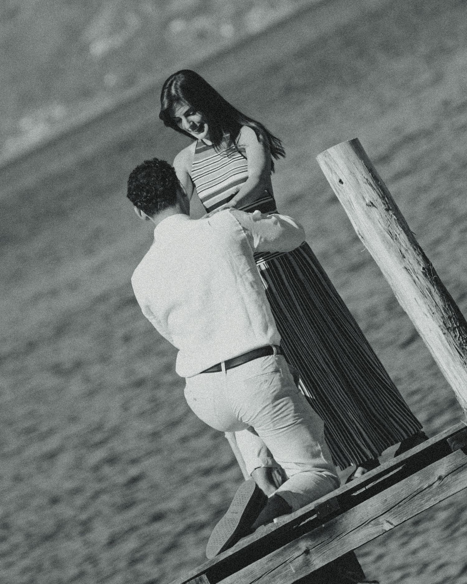 Black and white photo of a couple on a wooden dock by the water, woman in striped top smiling at man below.