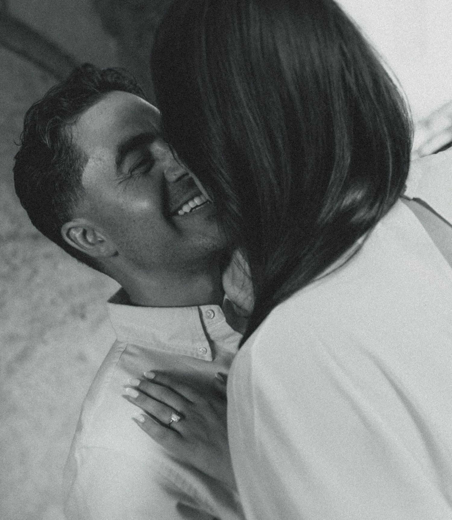 Black and white photo of a smiling man being embraced by a woman wearing an engagement ring.