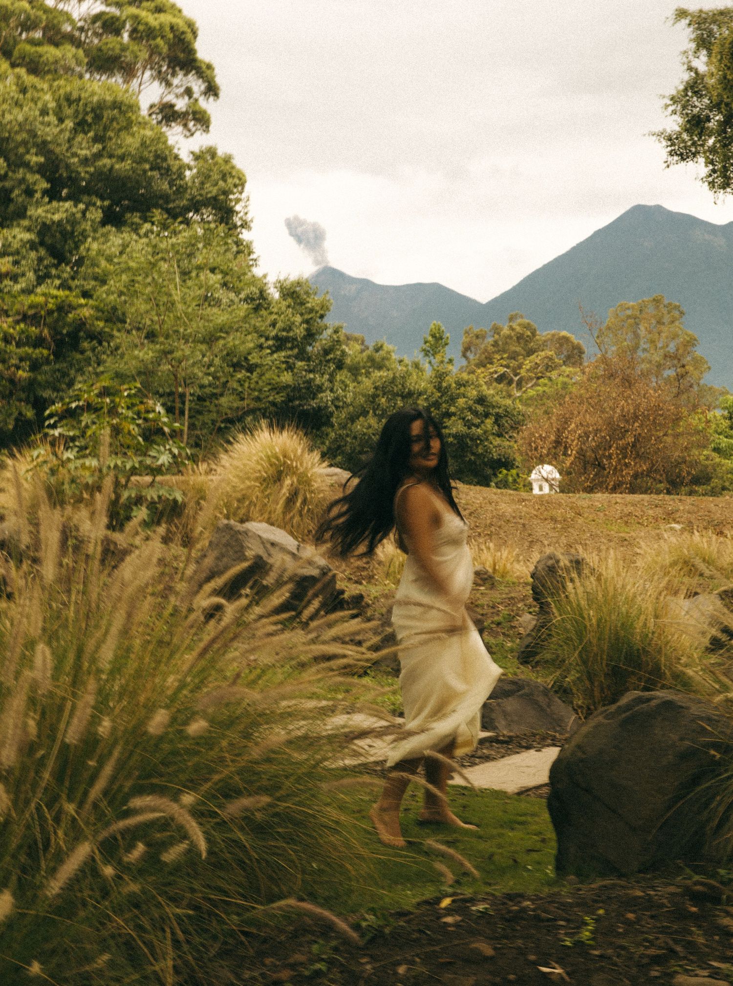 Woman in flowing cream dress walking through lush garden landscape with mountains in background.