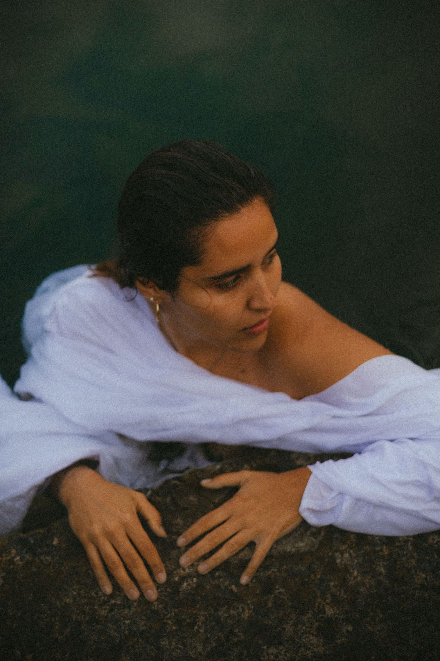 Woman in white off-shoulder blouse resting on rocks beside dark turquoise water with wet hair.