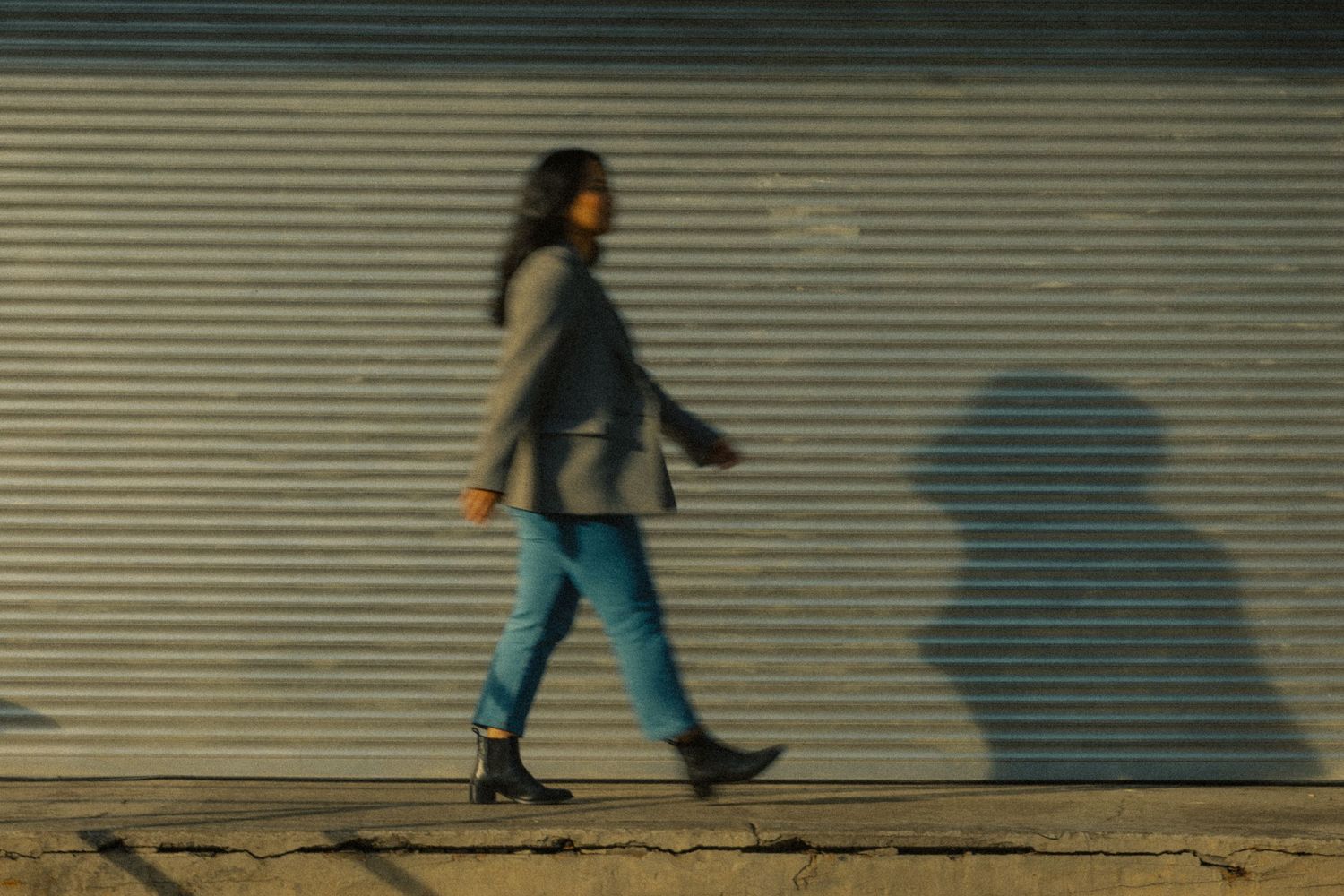 Woman walking past corrugated metal wall with shadow, motion blur street photography in urban setting.