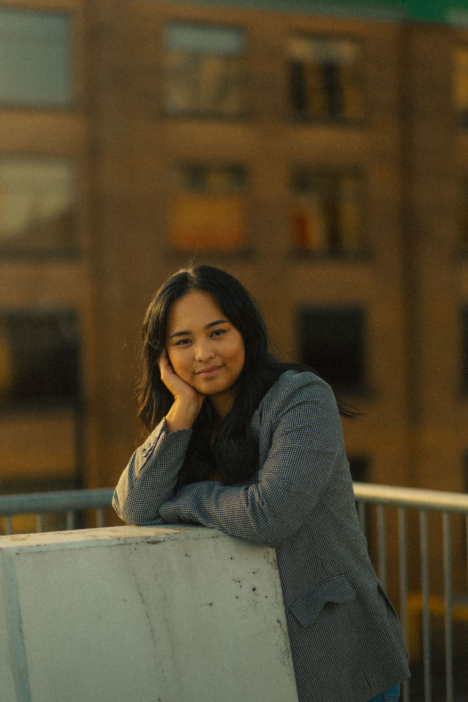 Young woman in gray blazer smiling, leaning on railing with blurred urban building in warm golden light.