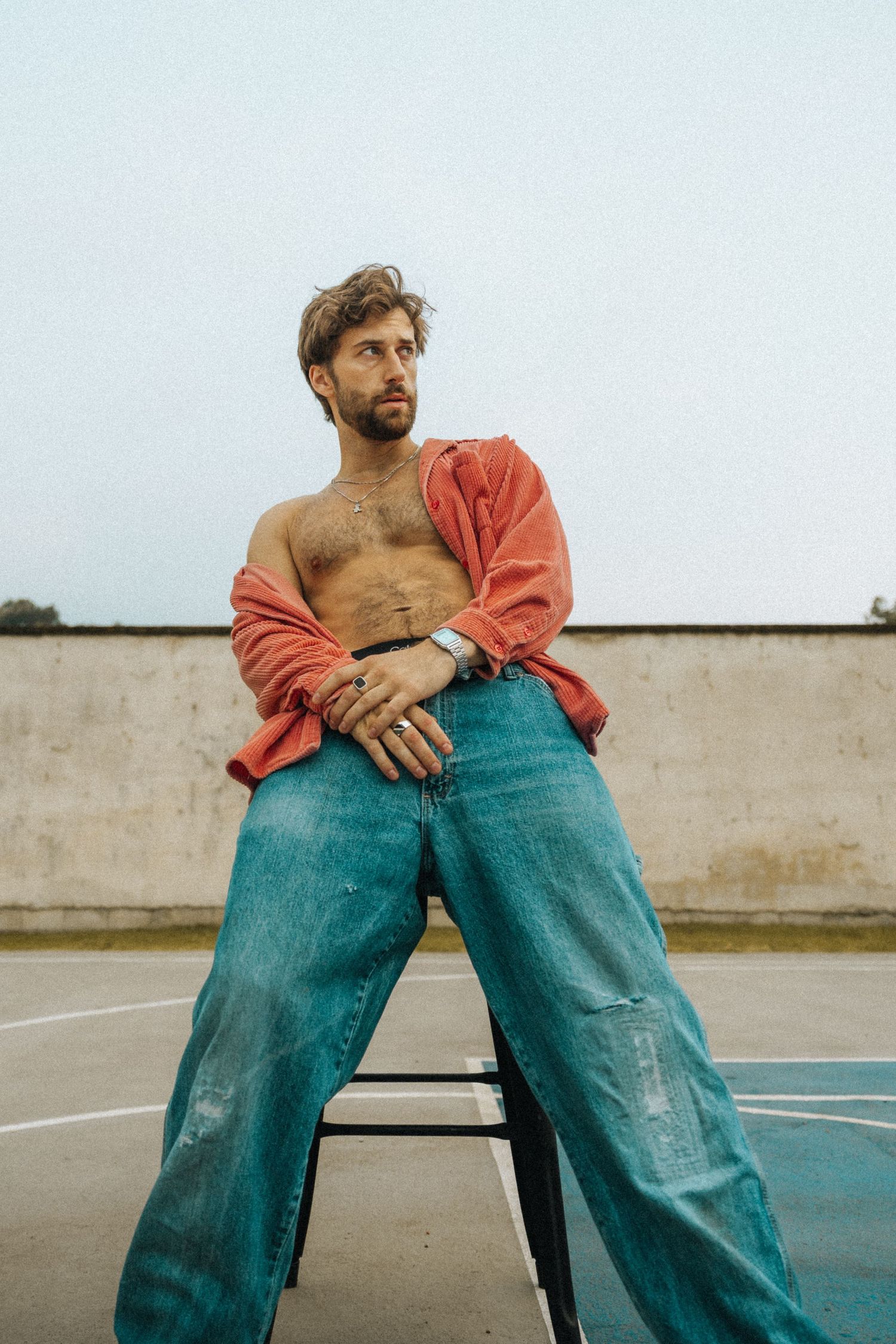Shirtless man in open red shirt and baggy jeans sits on chair outdoors on a basketball court.