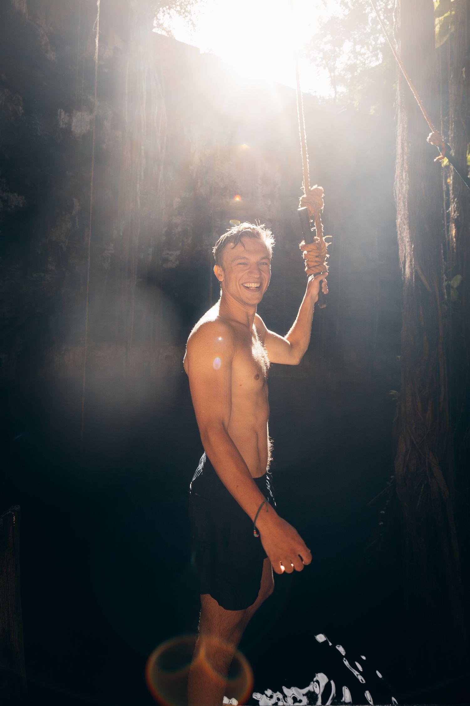Shirtless smiling man holds a rope swing in a sunlit jungle cenote with dramatic light rays.