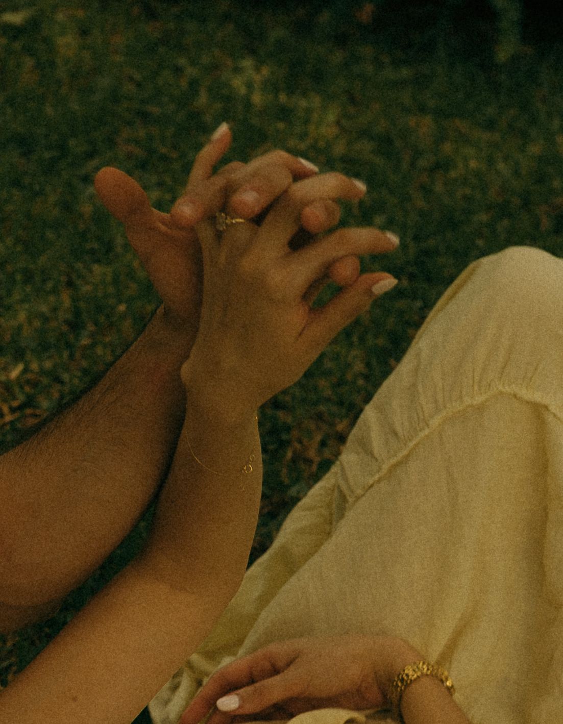 Close-up of couple holding hands with gold rings and bracelet on grass background.