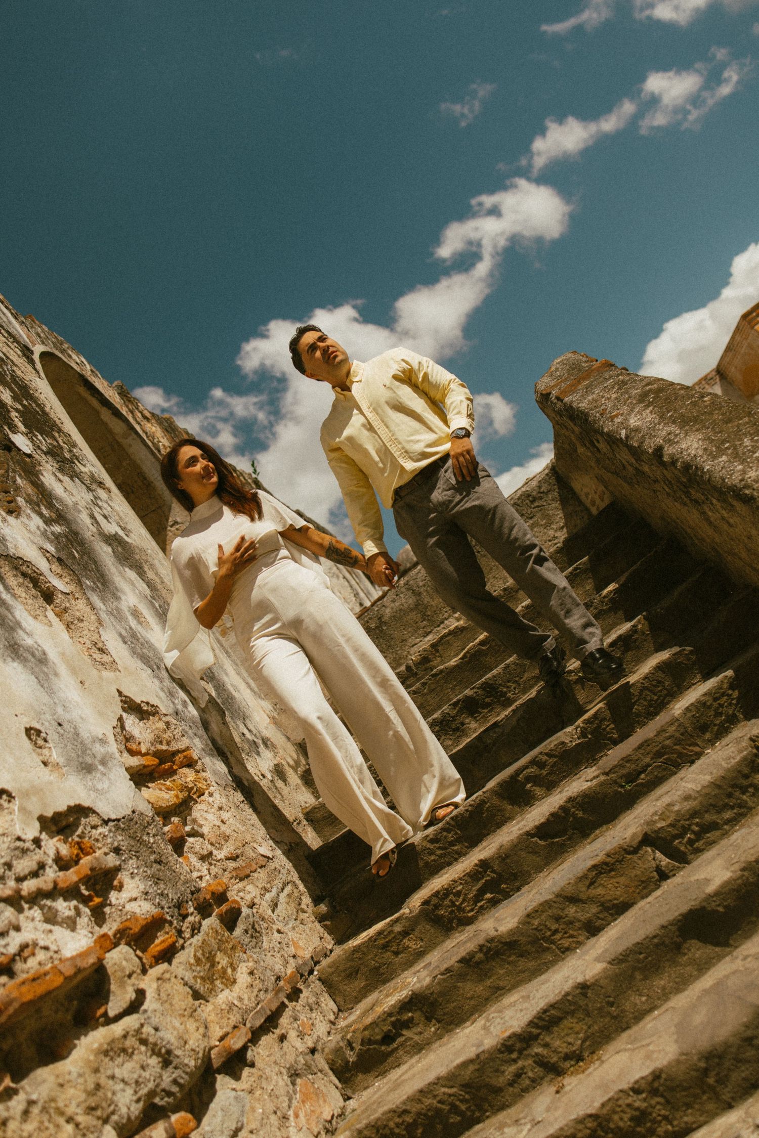 Couple in light, flowing outfits walk down weathered stone steps of an ancient building under a bright blue sky with white clouds.