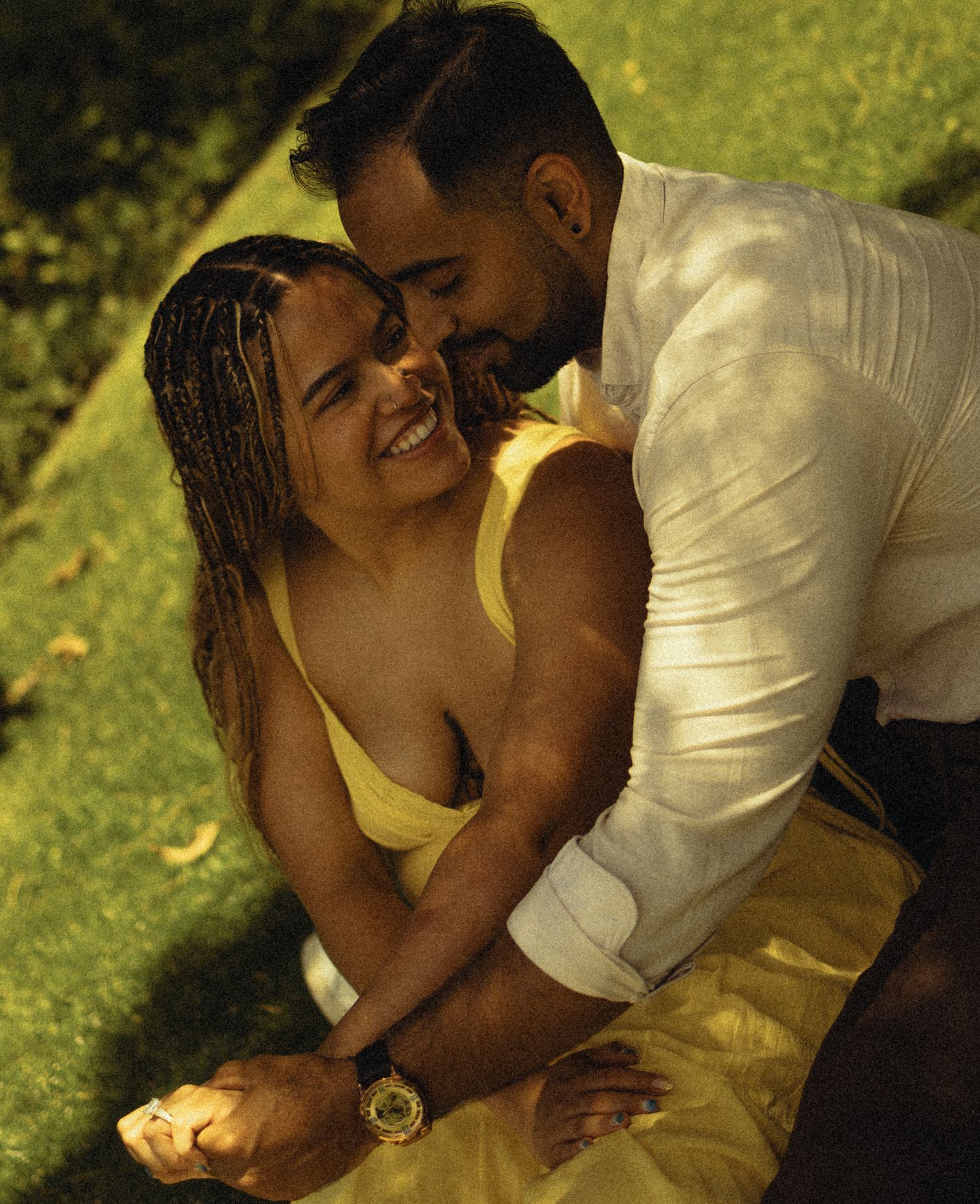 A joyful couple shares a romantic moment outdoors, with the woman in a yellow dress laughing as the man leans in close.
