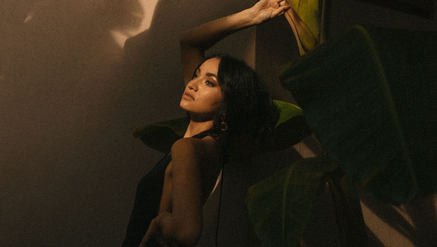 A woman poses dramatically against a wall with tropical leaves in warm, moody golden light.