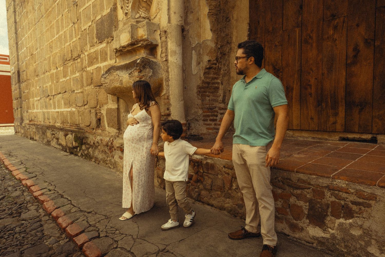 Pregnant woman in white dress holds hands with toddler son and husband on cobblestone street by historic stone wall.