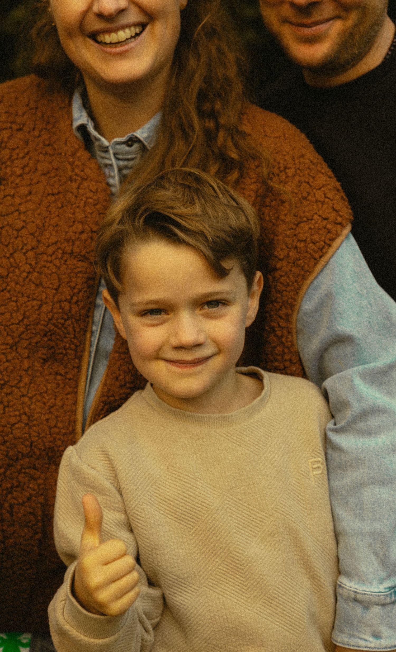 A young boy in a beige sweater gives a thumbs up while smiling, standing in front of two adults.