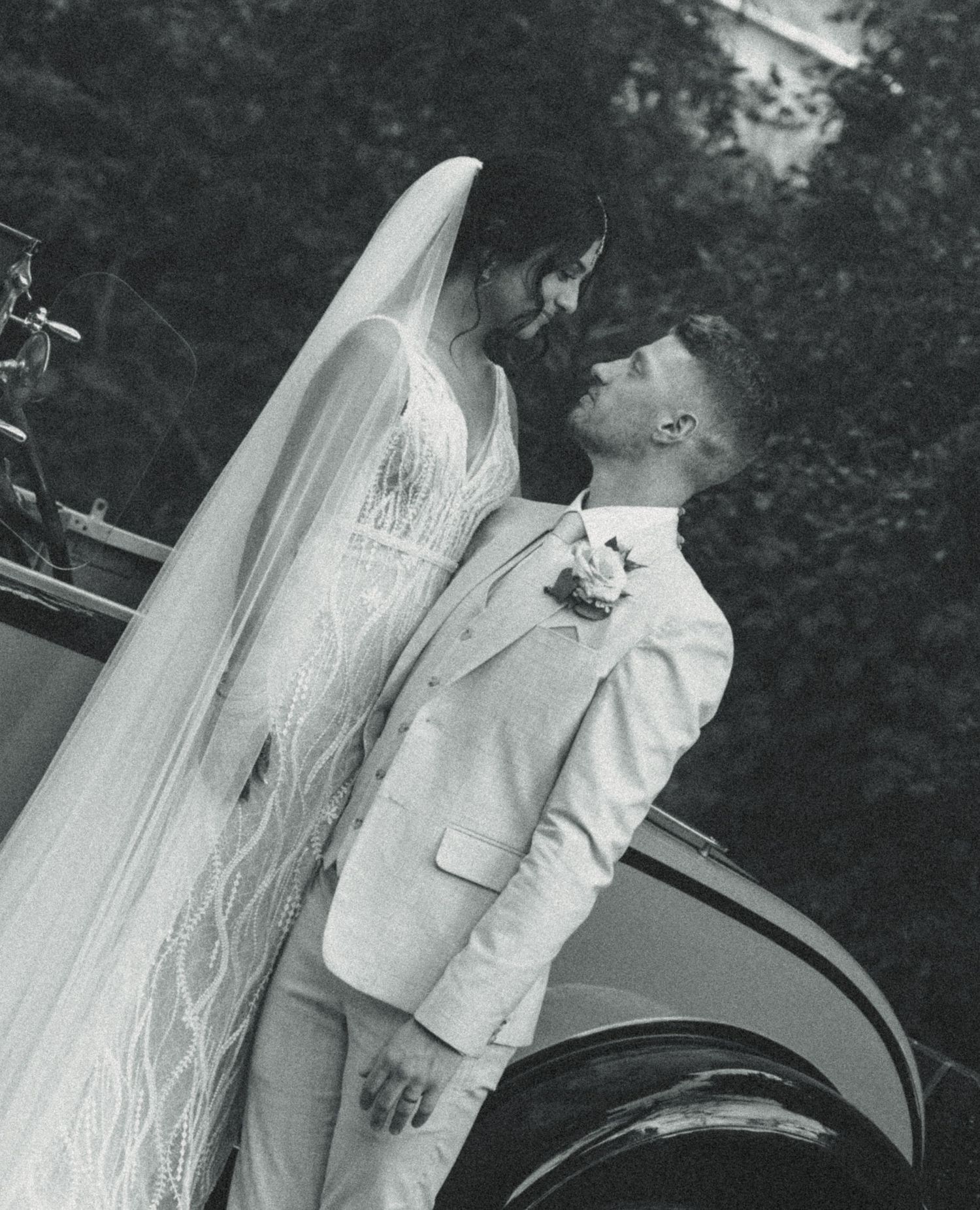 Black and white wedding photo of bride in lace gown with veil and groom in white suit beside vintage car.