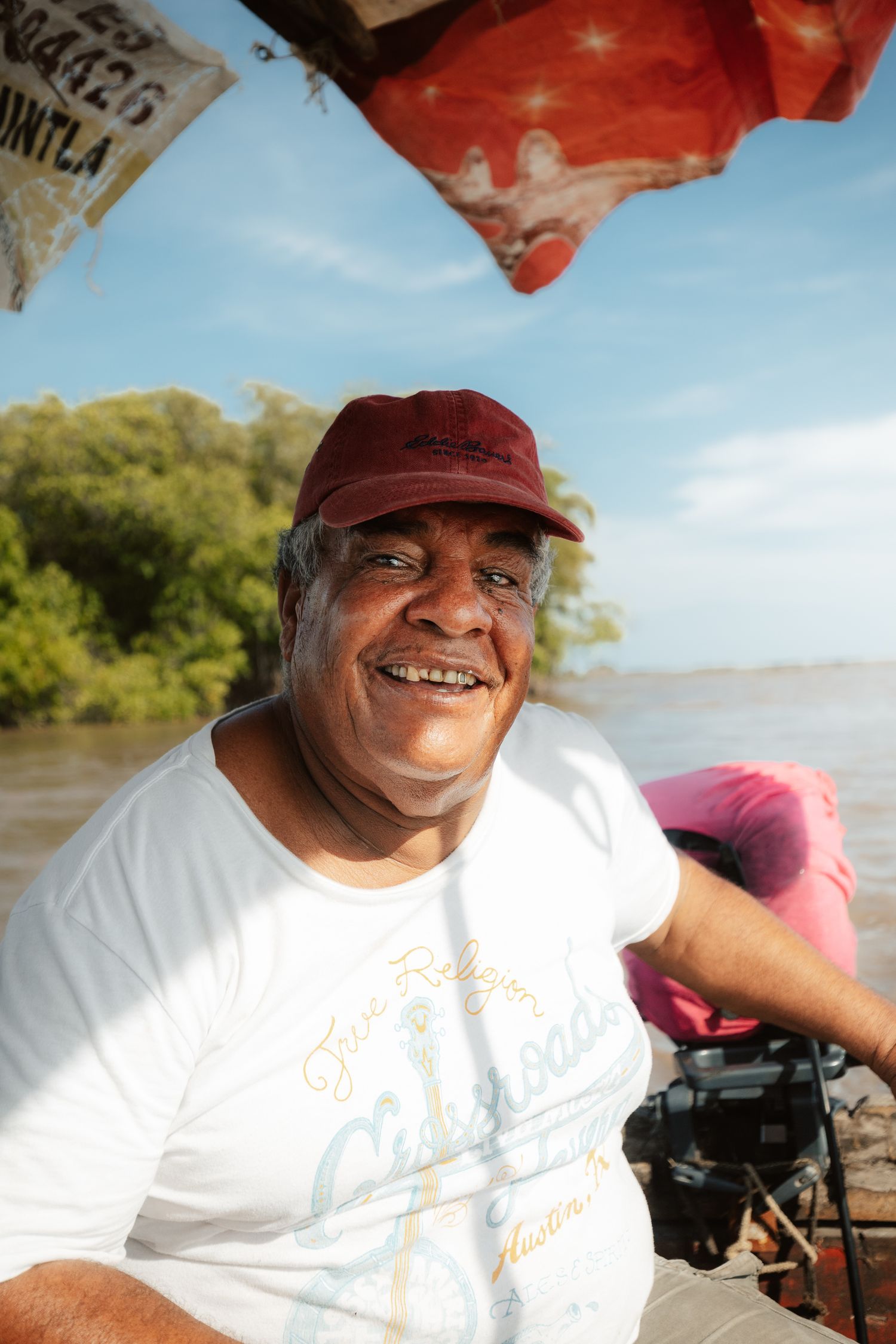 Smiling man in red cap and white t-shirt sitting on a boat with river and green trees in background.