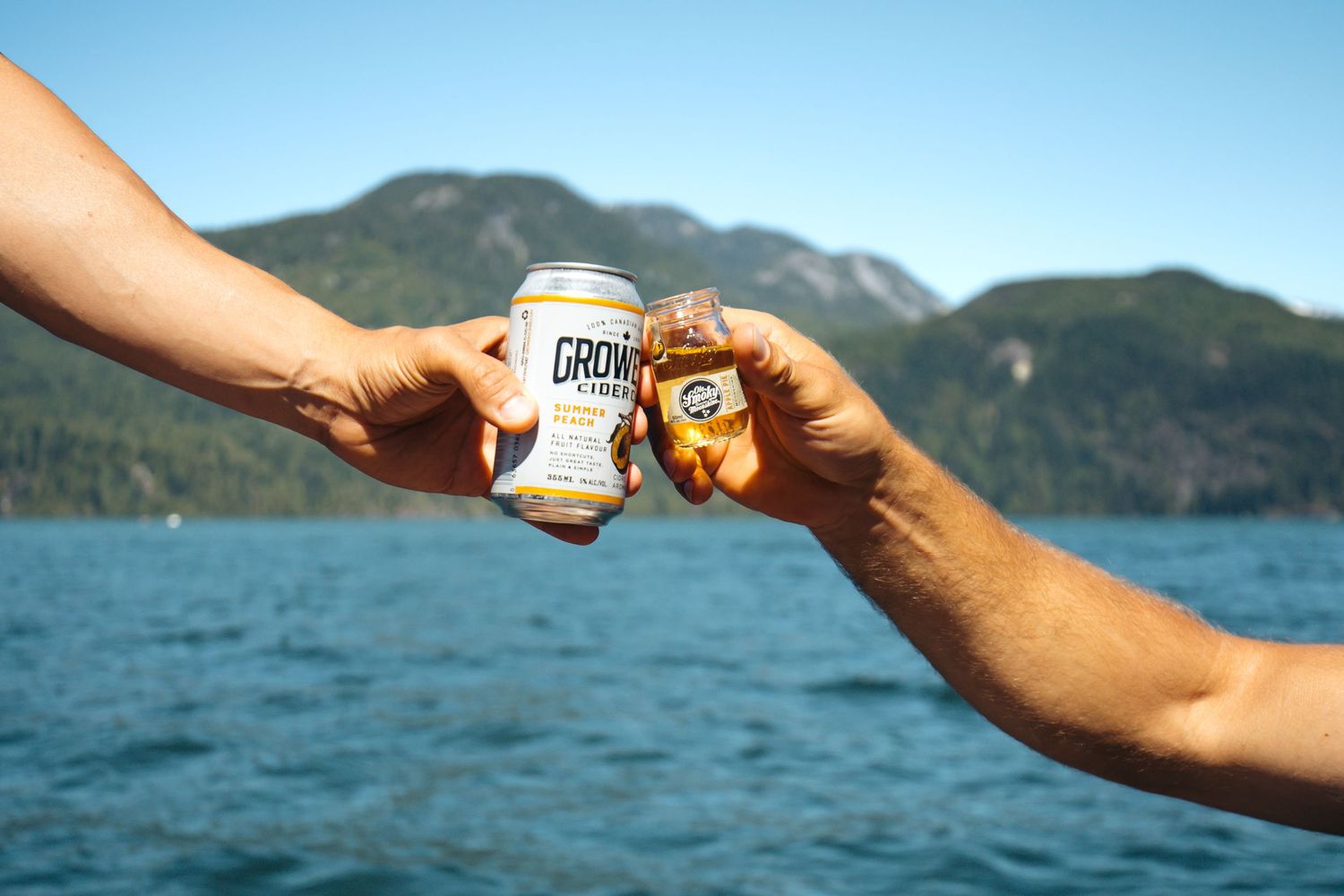 Two hands toasting with Groin beer cans over a scenic mountain lake on a sunny day.