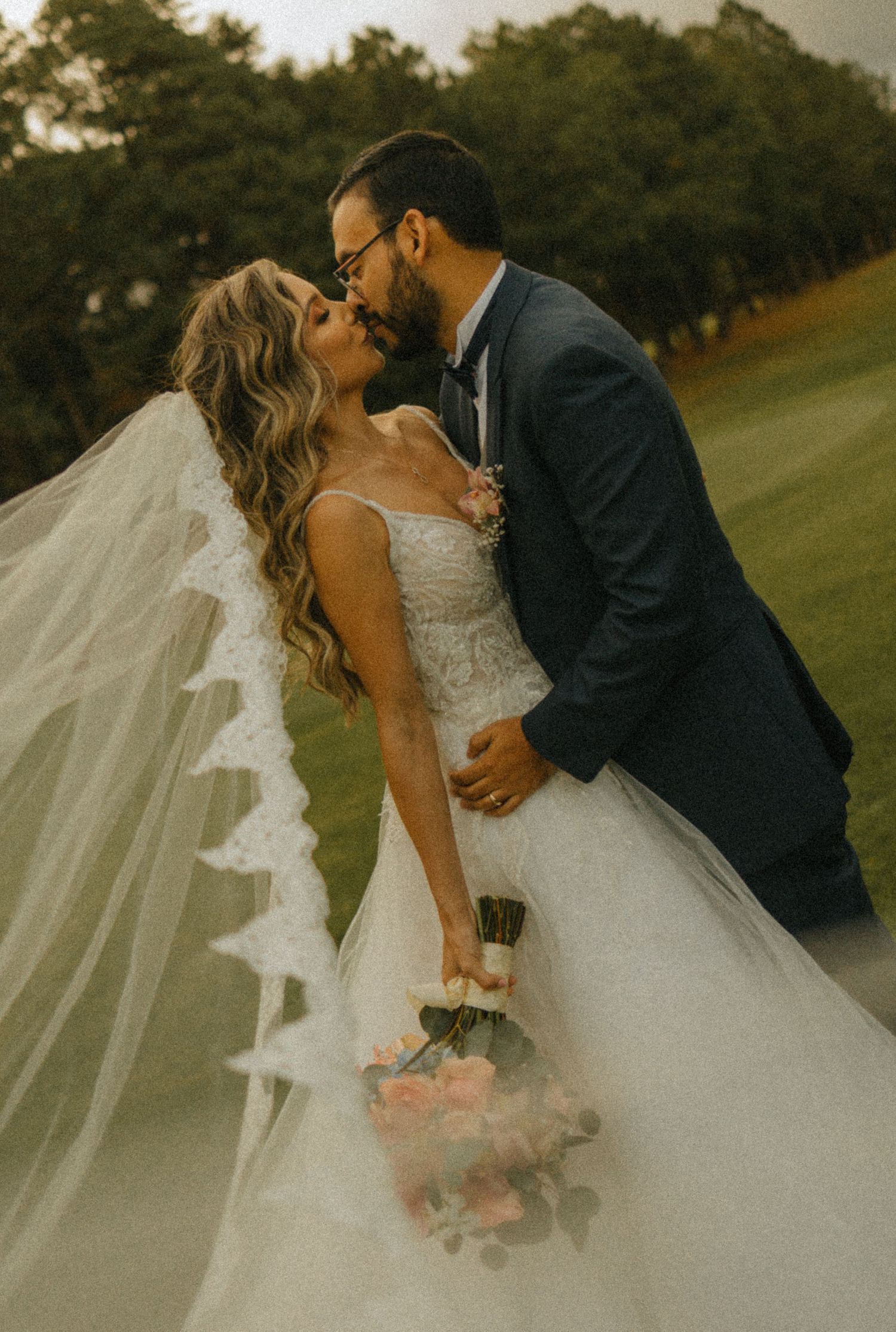 A bride and groom share a romantic kiss outdoors, her lace veil flowing in the wind as she holds a pink bouquet.