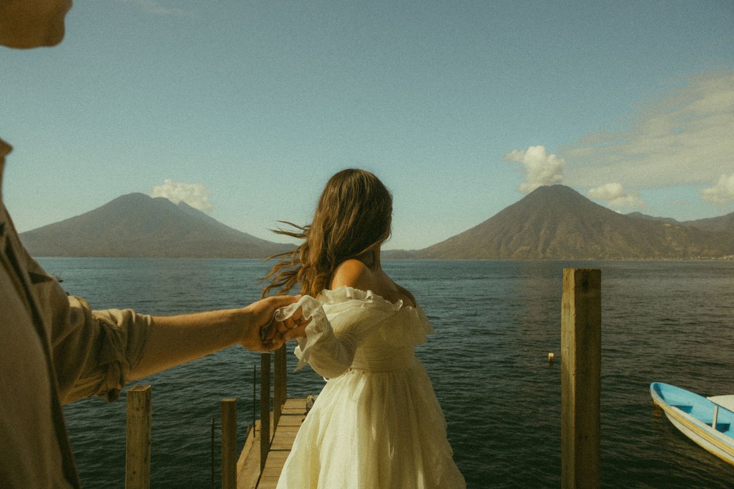 Woman in white dress on wooden dock with volcanic mountains and blue lake in background.