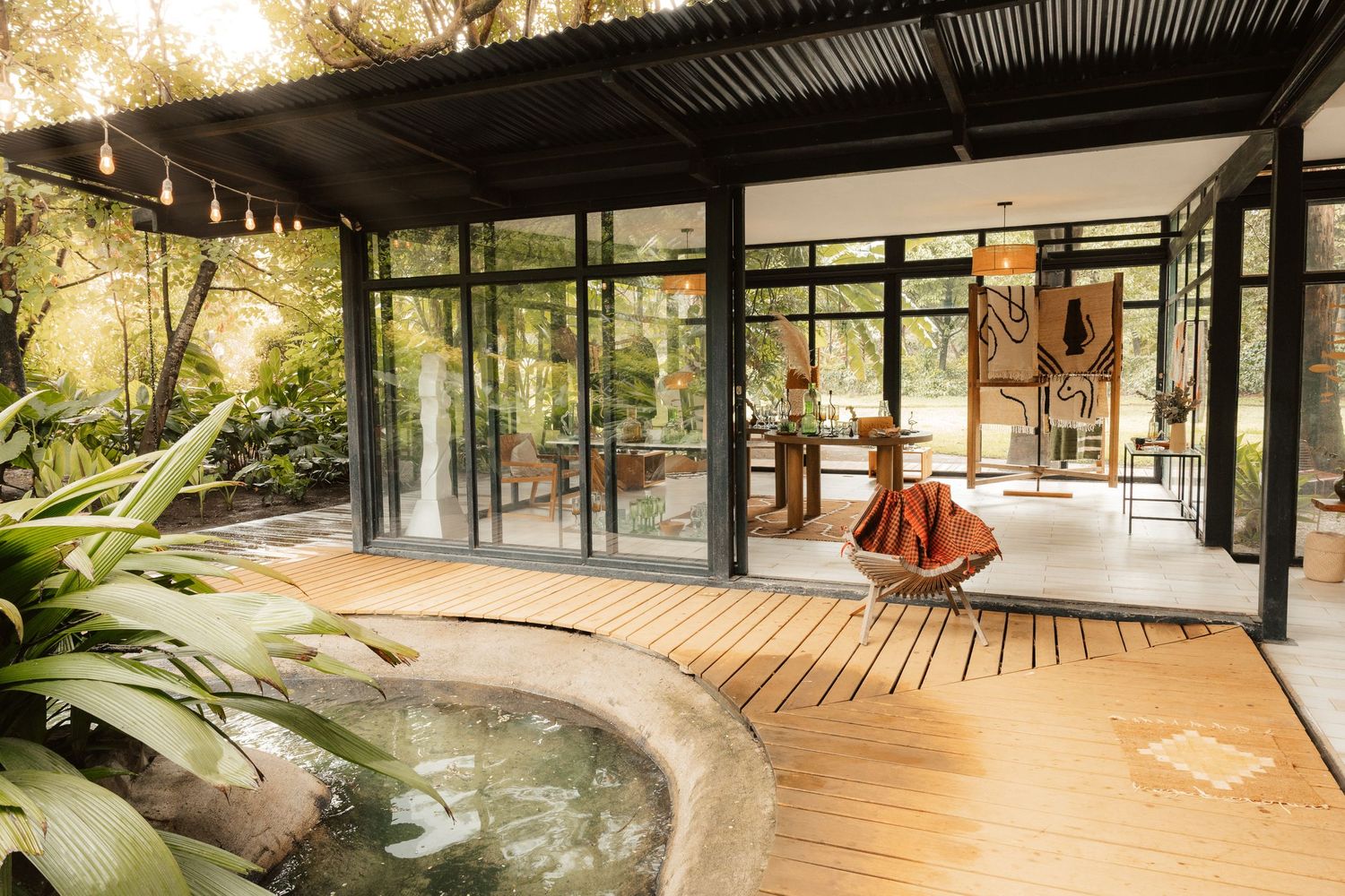 Modern glass pavilion with wood deck, circular pool, and lush tropical garden surroundings in natural light.
