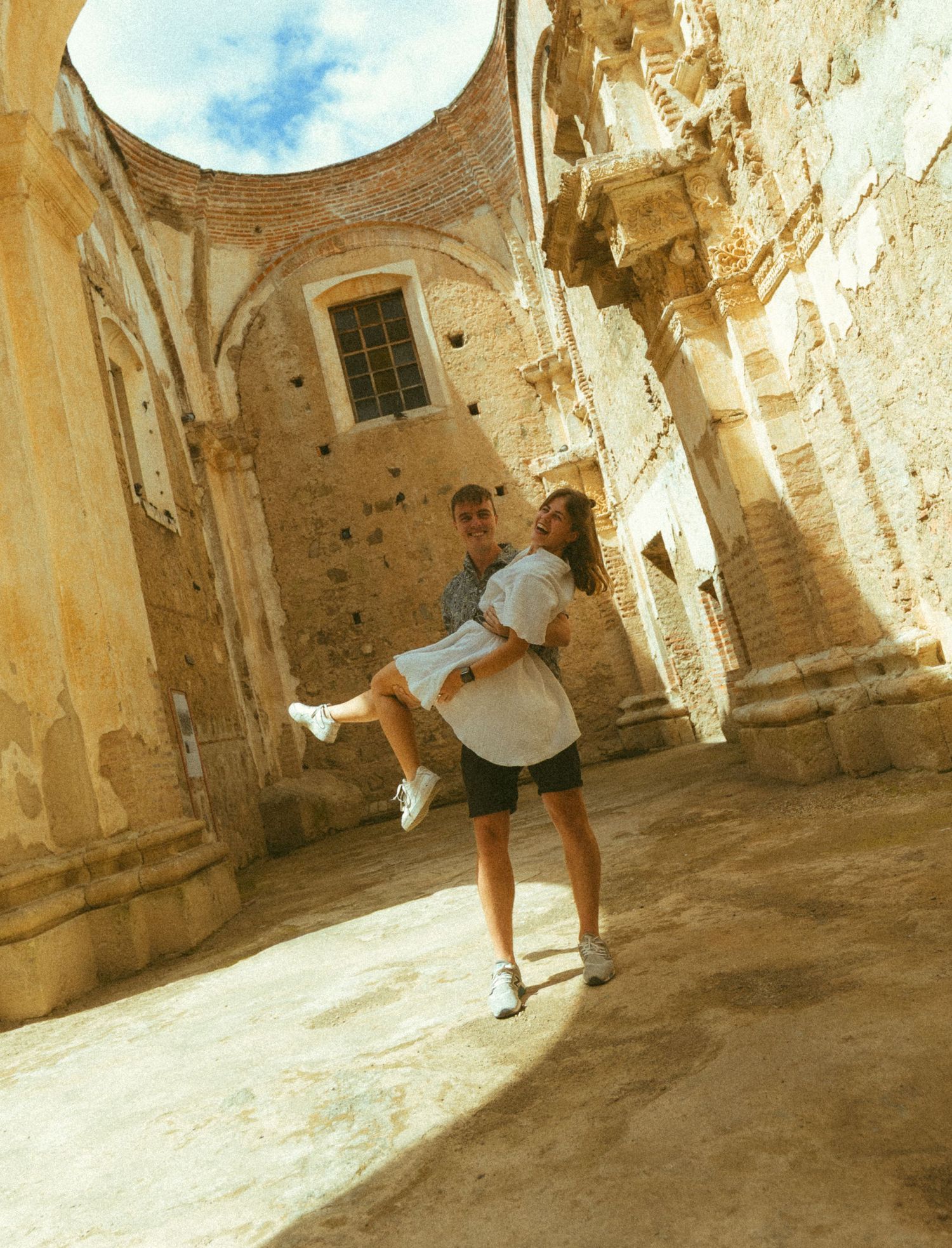 Couple posing romantically in ancient stone ruins, man lifting woman in white dress, historic architecture background.