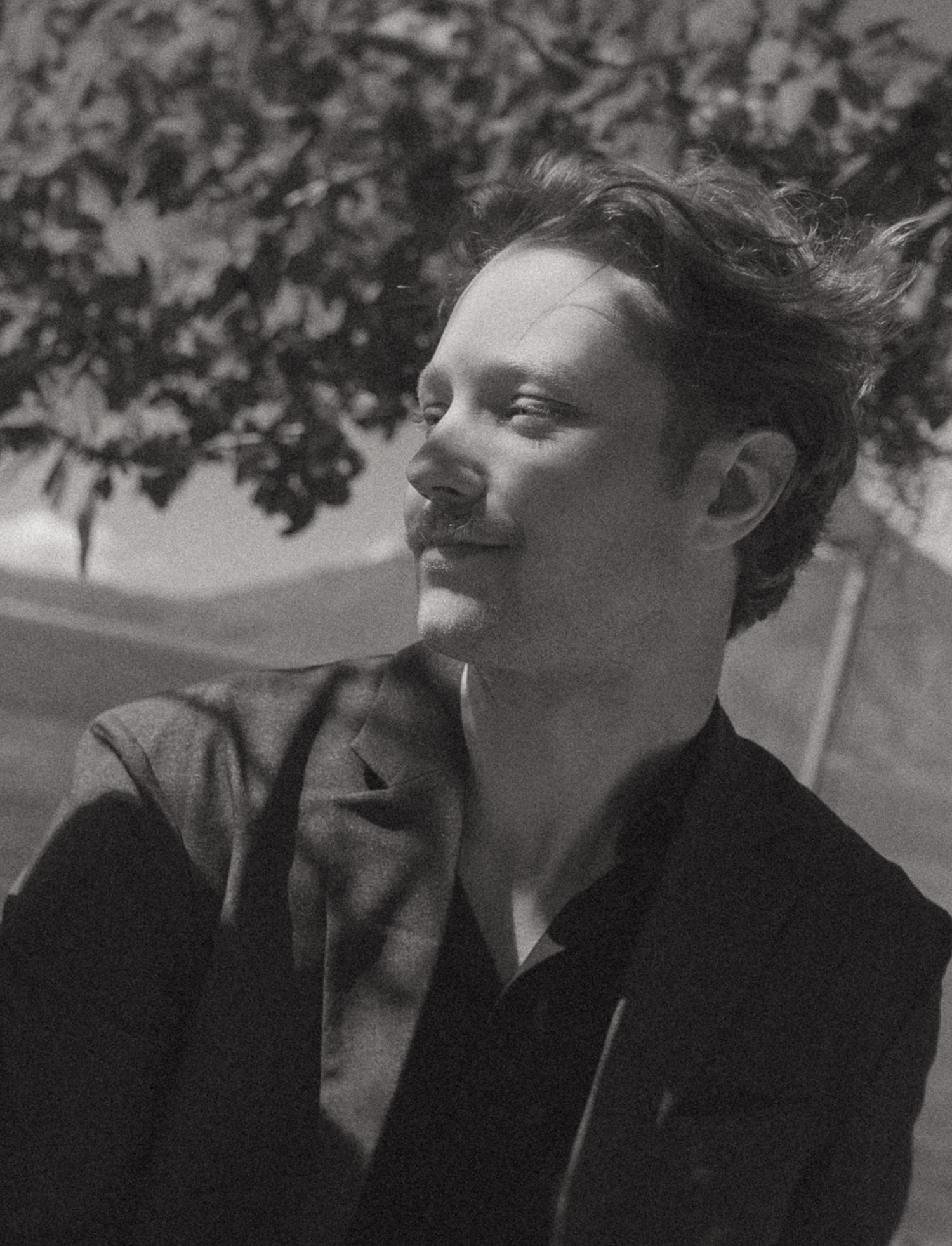 Black and white portrait of a young man with mustache sitting outdoors under a tree.