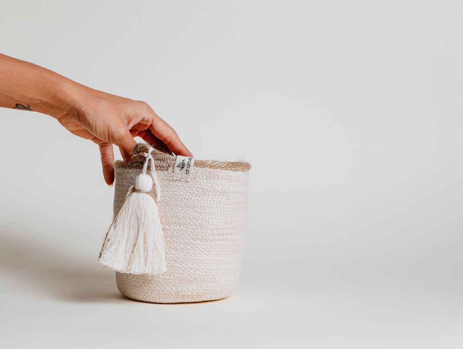 Hand holding a woven rope basket with white tassel charm against a light gray background.