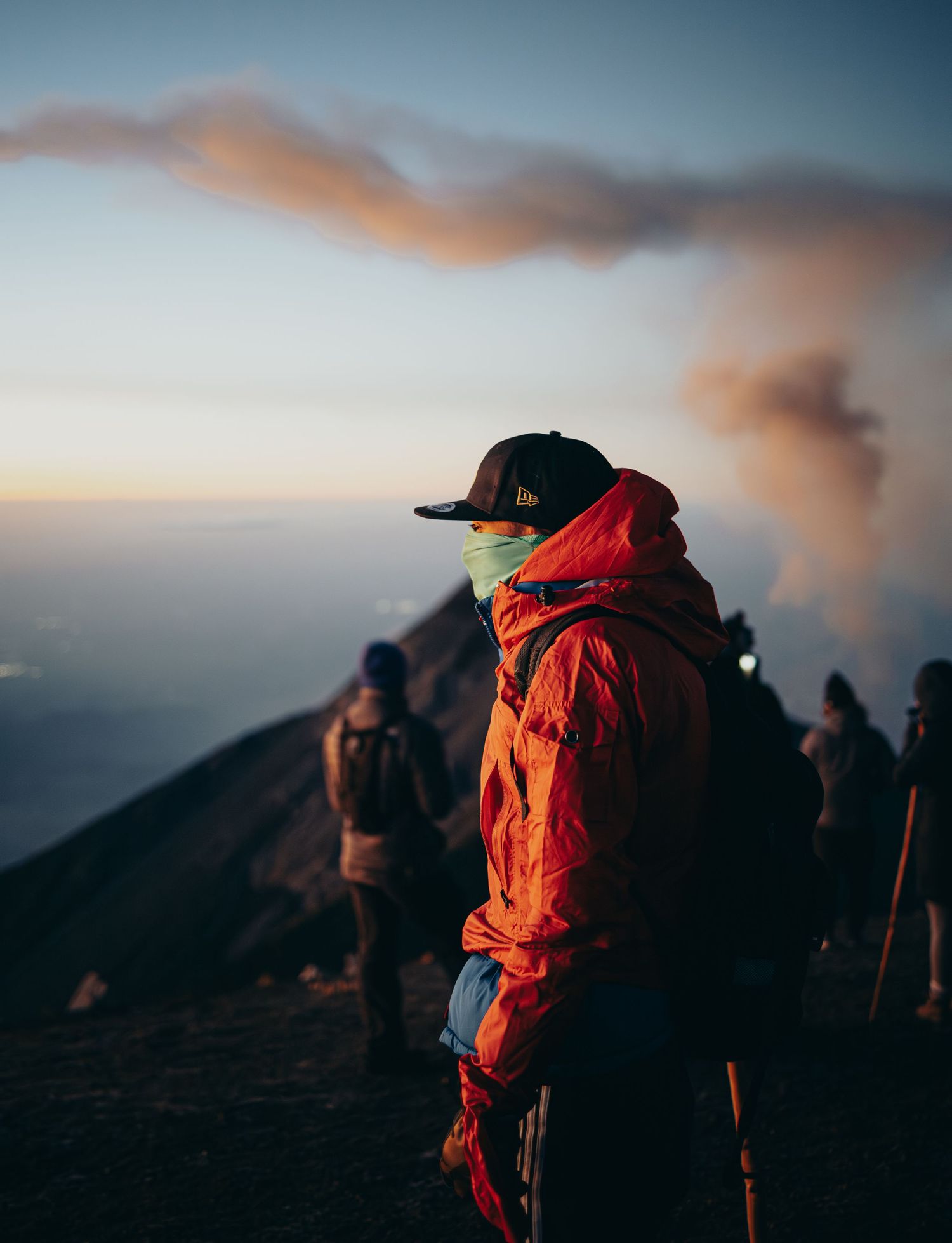 Hiker in orange jacket and face mask stands on volcanic ridge at dusk with smoke rising in background.