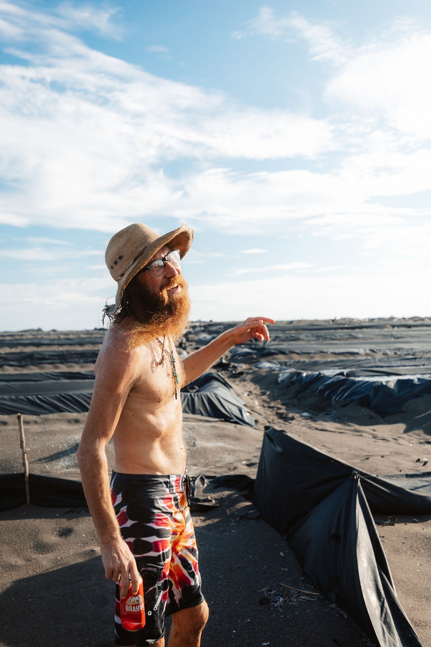 Shirtless bearded man in straw hat and board shorts pointing outdoors near black tarp-covered ground under blue sky.