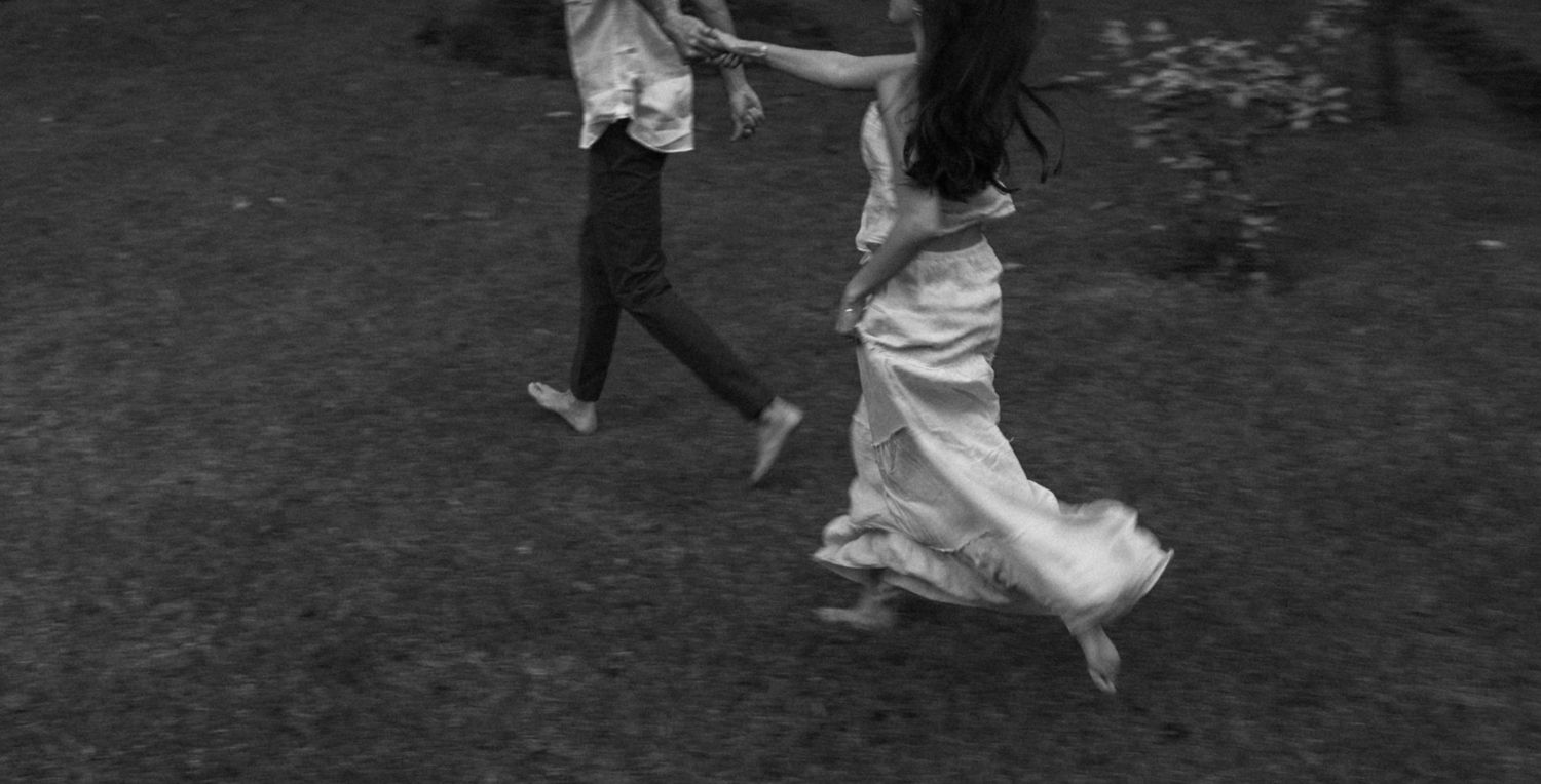 Black and white photo of two people dancing joyfully outdoors, woman in flowing white dress, captured in motion.