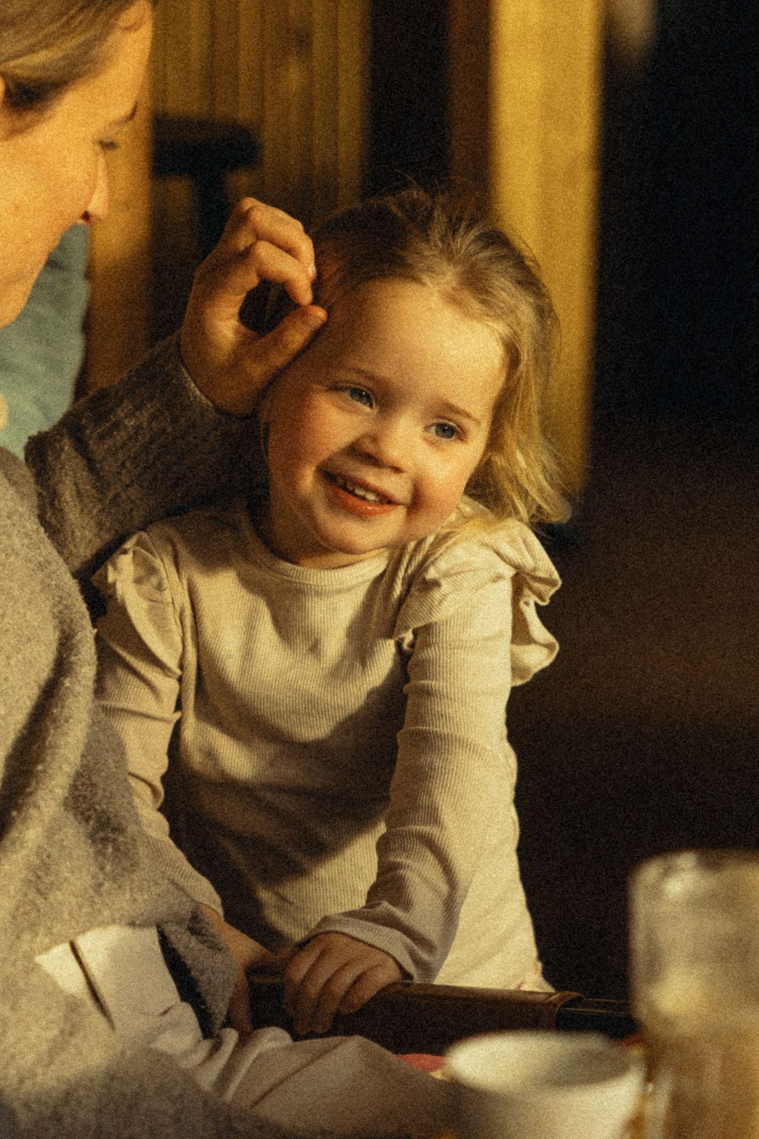 A smiling toddler girl in a white top enjoys a warm moment while an adult gently fixes her hair.