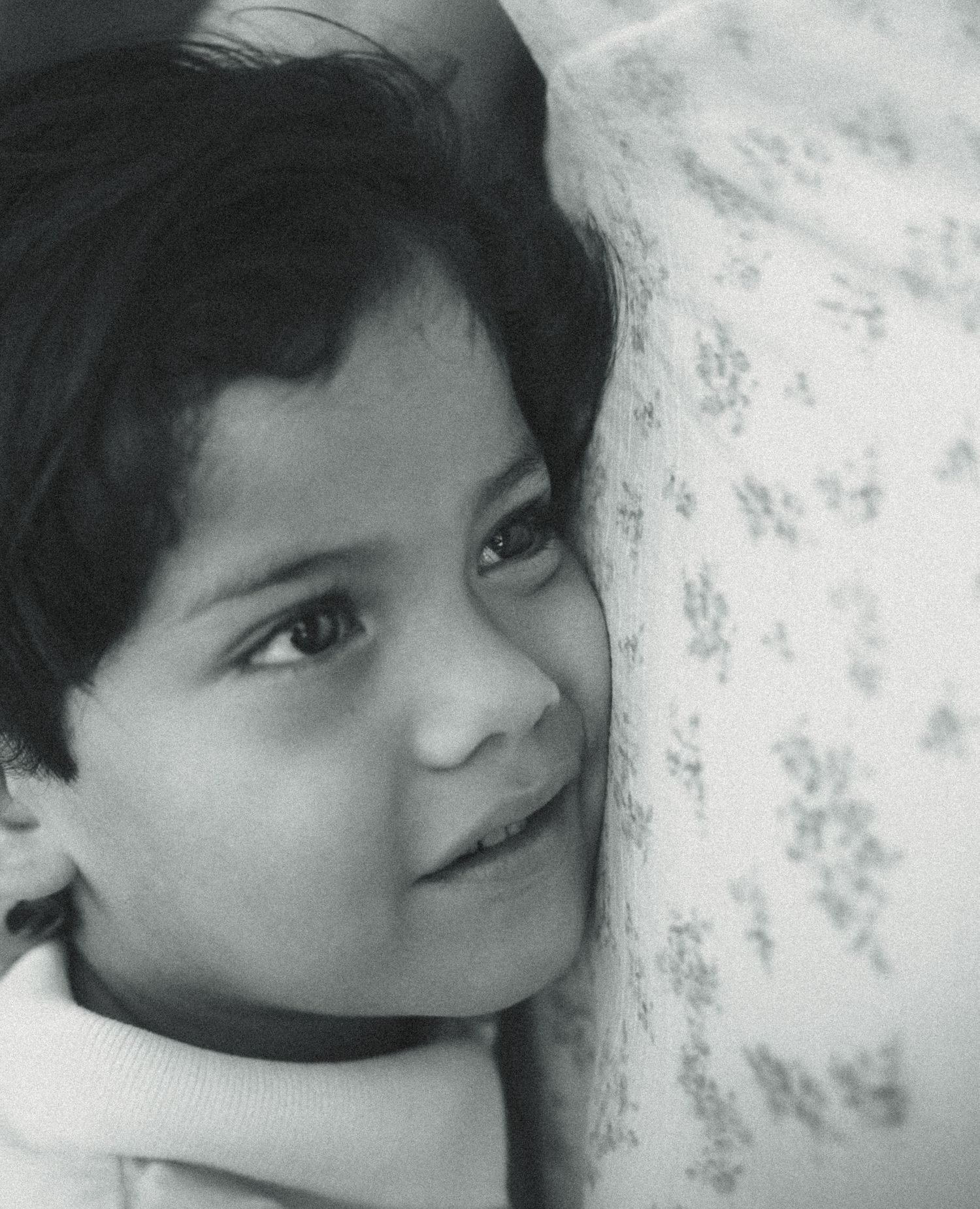Black and white portrait of a young child smiling shyly, resting cheek against a floral patterned surface.