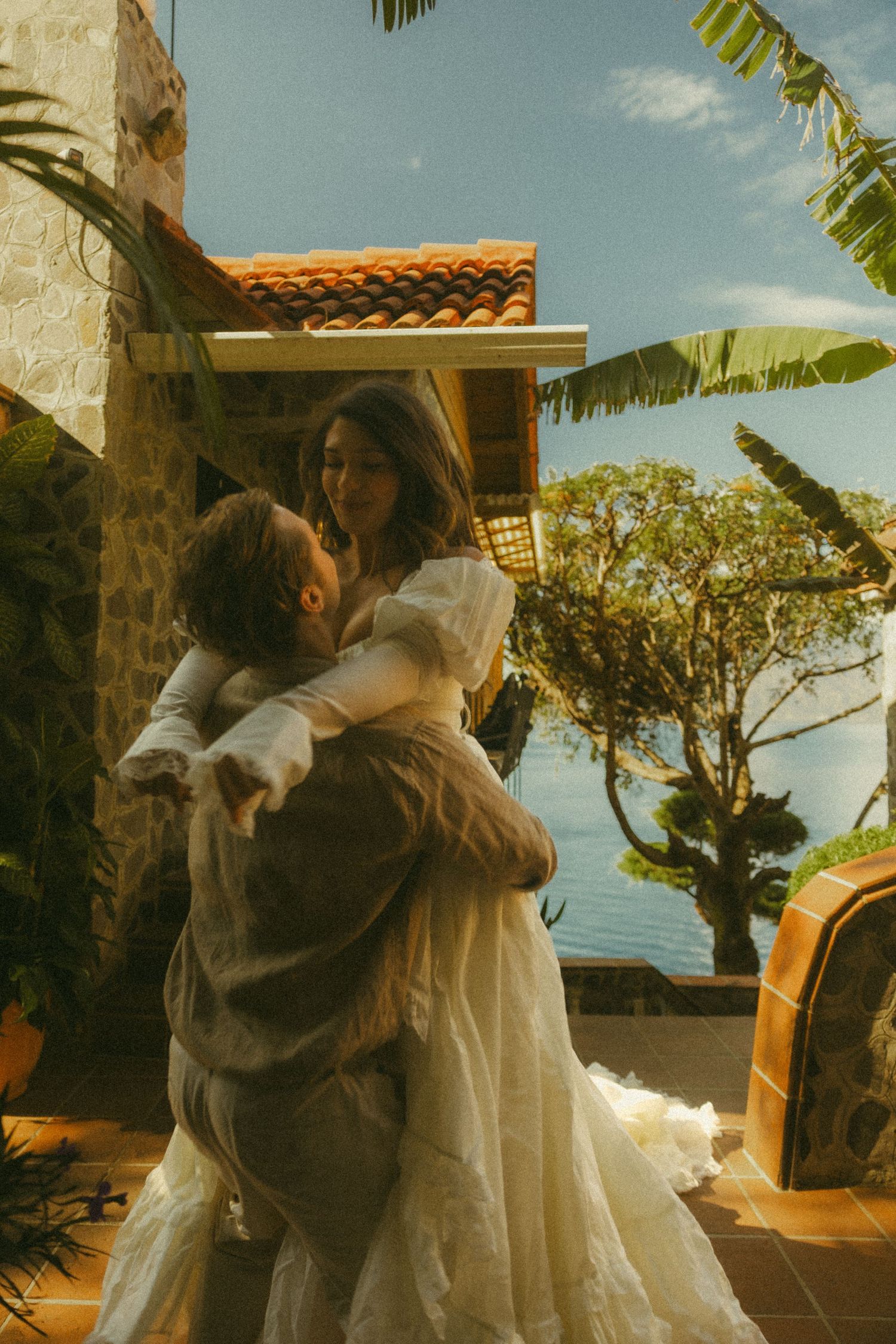 Groom lifting bride in white wedding dress outside a Mediterranean villa with ocean views and tropical trees.