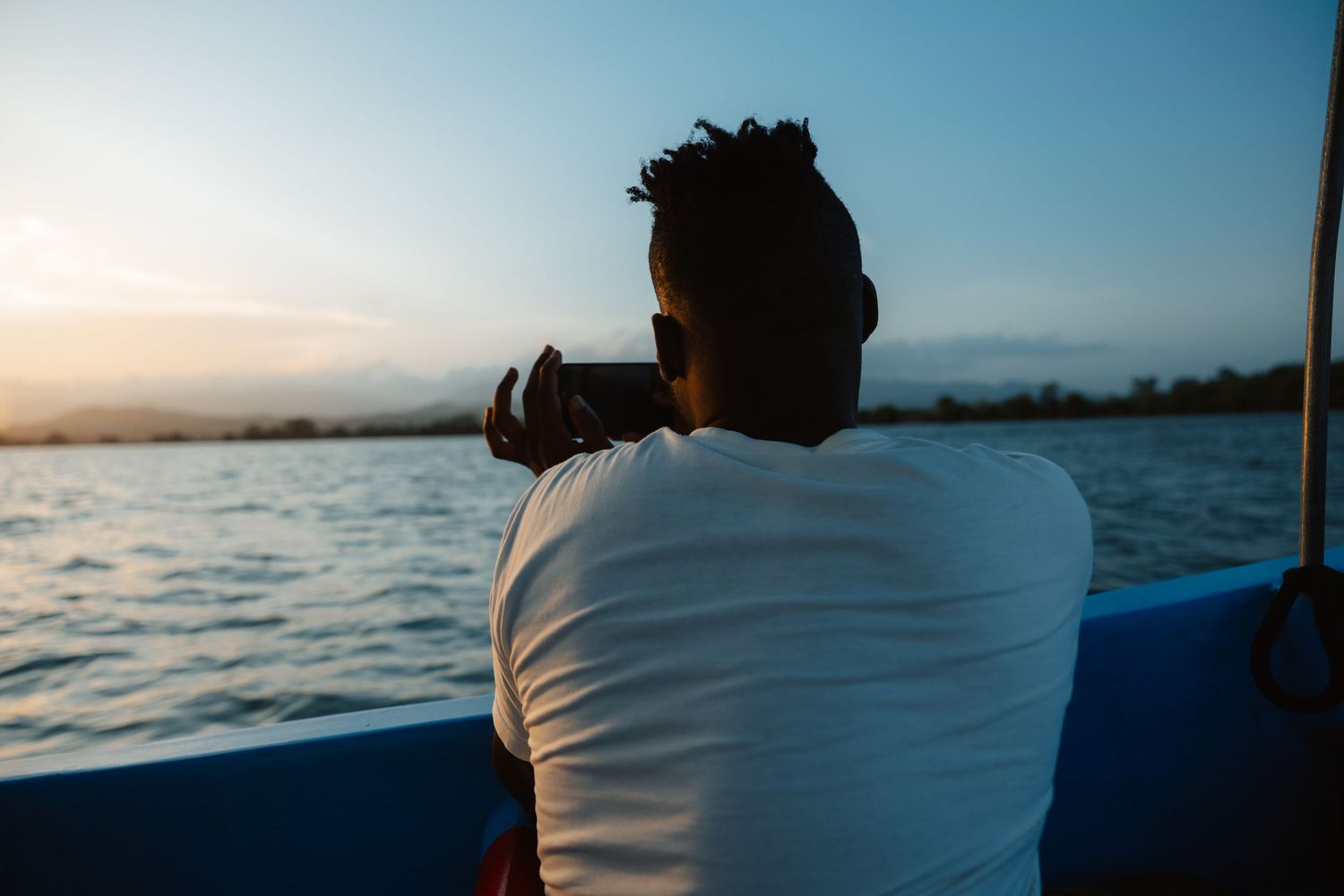 Man in white t-shirt photographing a serene sunset over calm water from a boat.