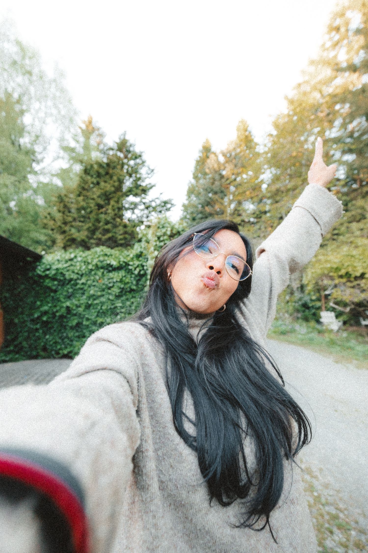 Young Asian woman with long dark hair takes a selfie outdoors, wearing glasses and a gray sweater, pointing up.