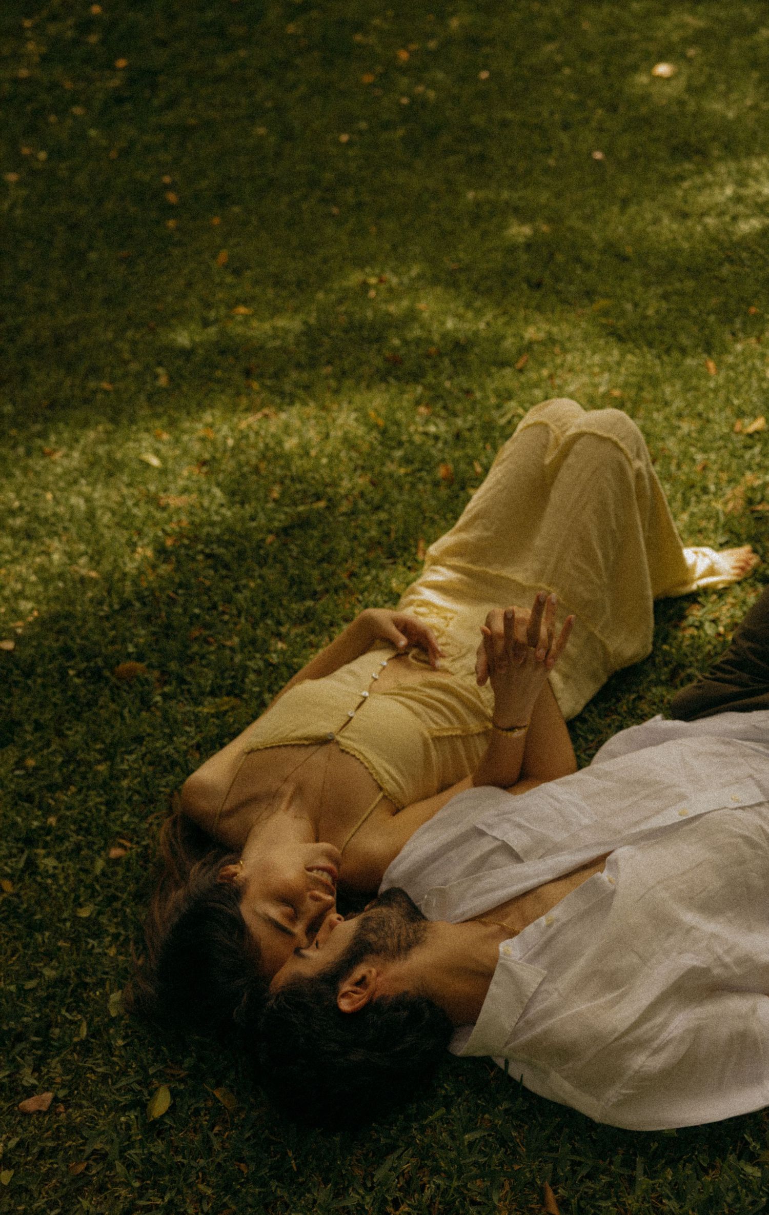 A couple lying on green grass, embracing romantically, wearing light yellow and white summer outfits.