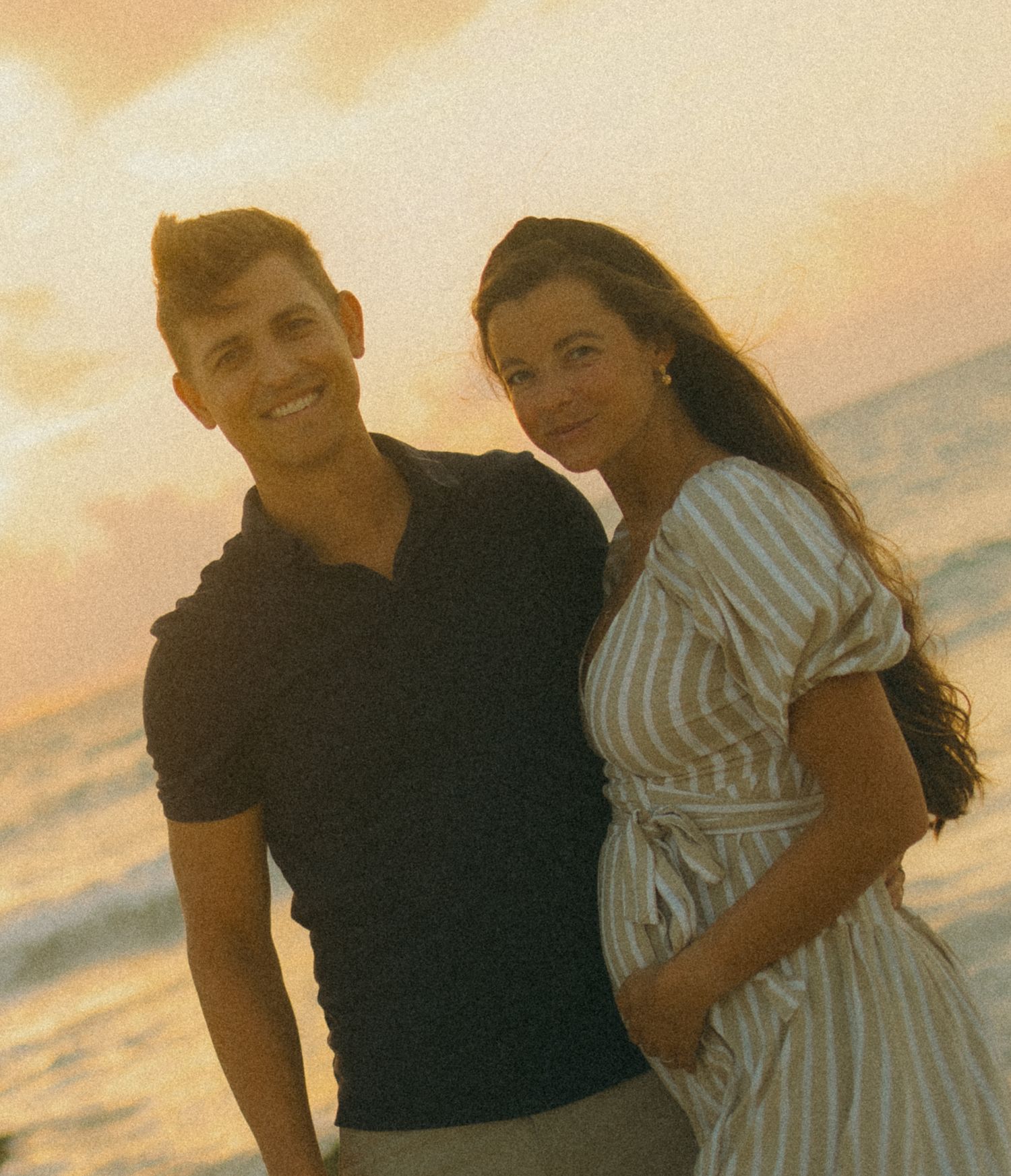 A smiling couple poses romantically on a beach at sunset, man in navy polo and woman in striped dress.