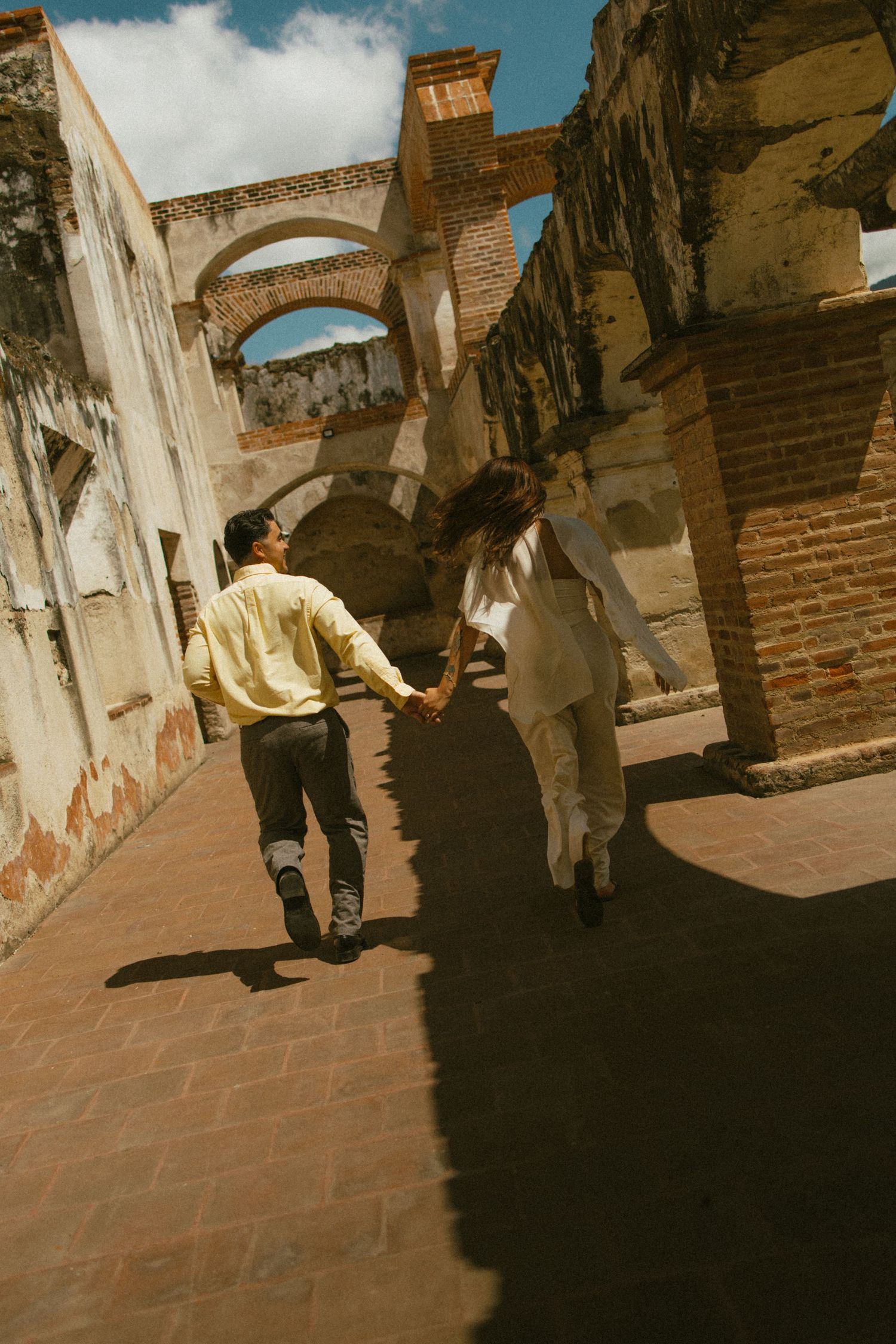 Couple holding hands and running through the sunlit brick arches of historic colonial ruins on a warm, sunny day.