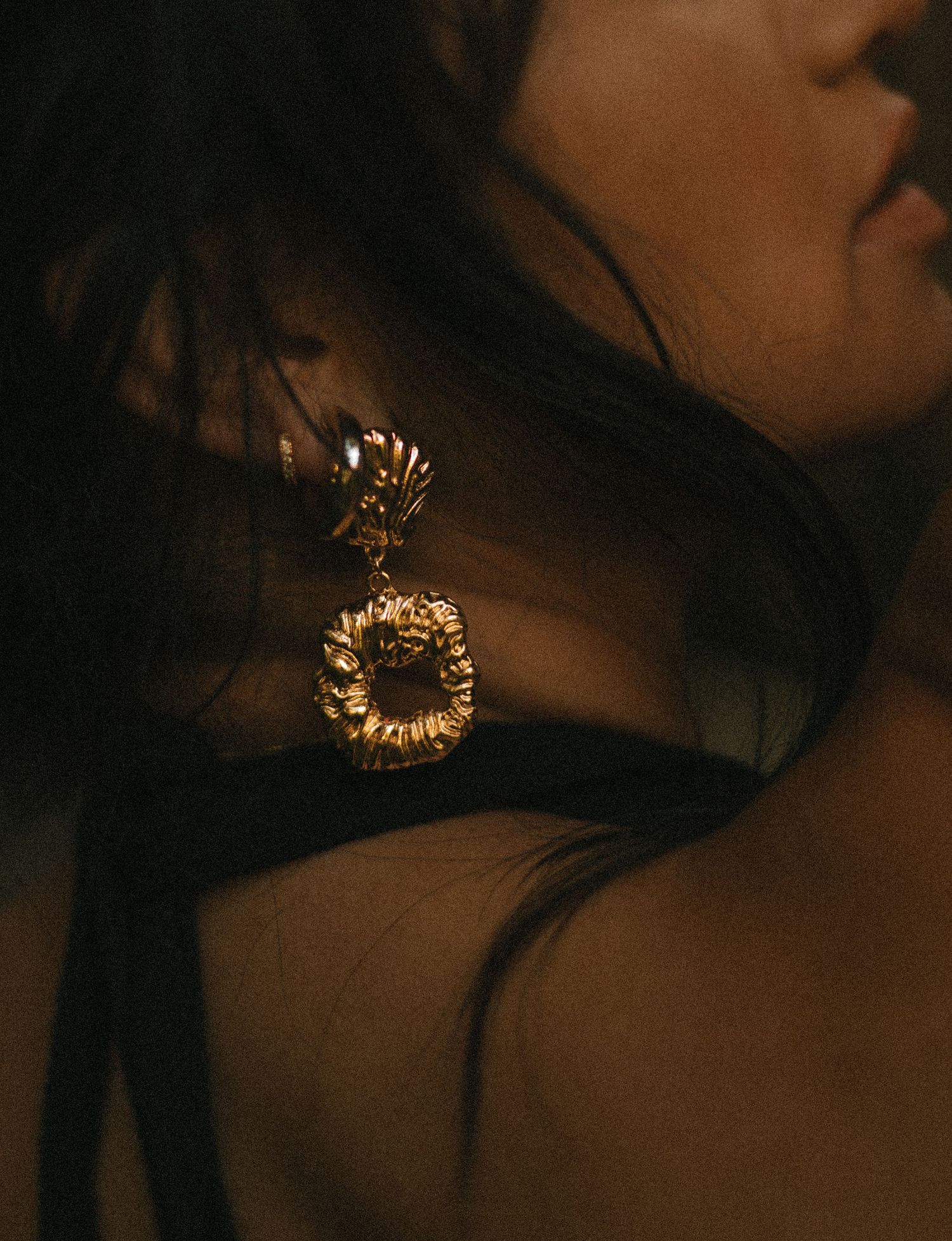 Woman wearing ornate gold drop earrings with textured circular pendant, photographed in warm moody lighting.