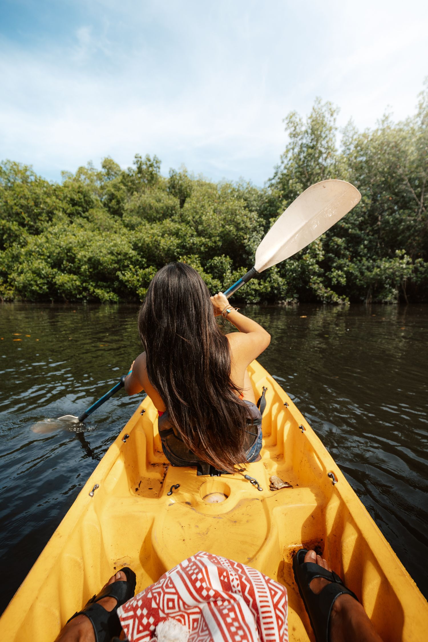 Woman kayaking in a yellow kayak through a lush green mangrove river, holding a paddle on a cloudy day.