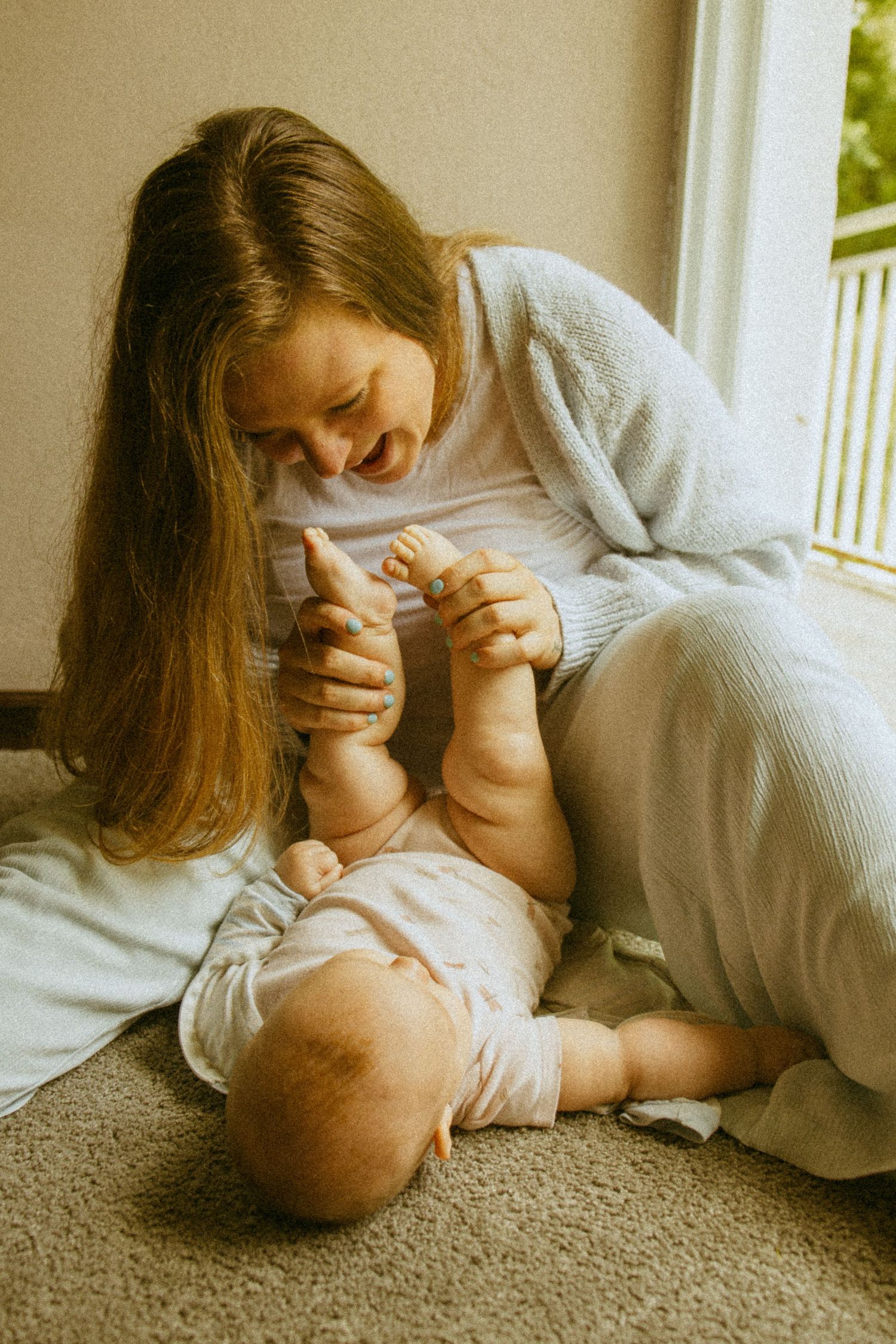 A young mother sitting on the floor, joyfully playing with her baby's feet while the infant lies on a soft carpet.