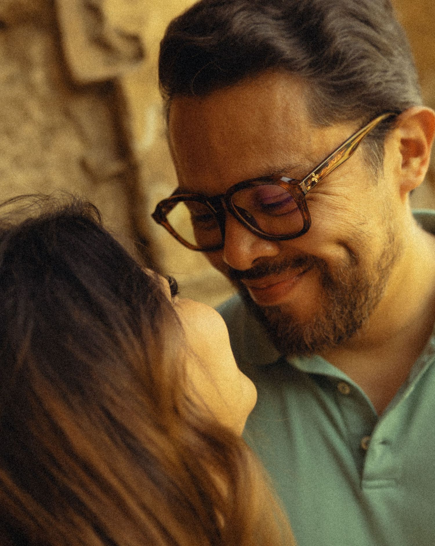 A smiling bearded man with glasses gazes down at a woman with long hair in a warm, intimate moment.