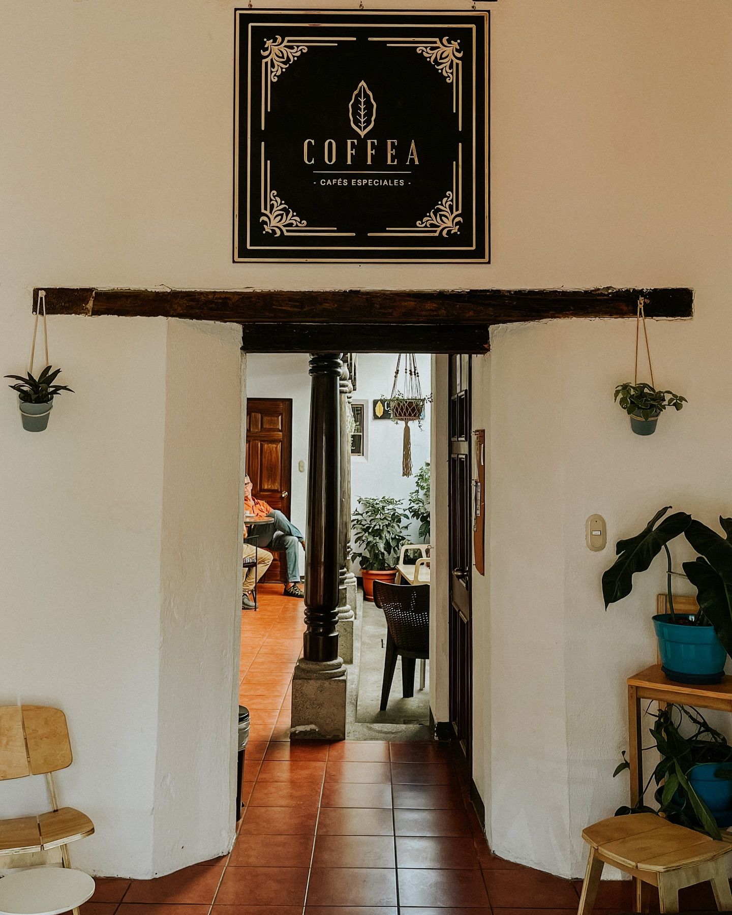 Entrance to Coffea Cafes Especiales coffee shop with rustic wooden beam, hanging plants, and terracotta tile floors.