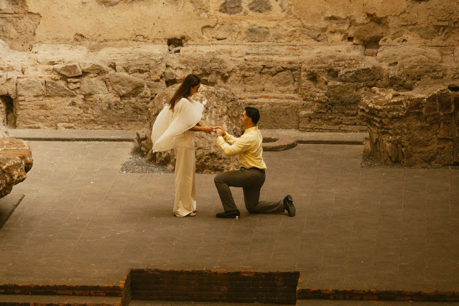 Man proposes on one knee to woman in white dress at ancient Roman ruins, a romantic engagement moment.