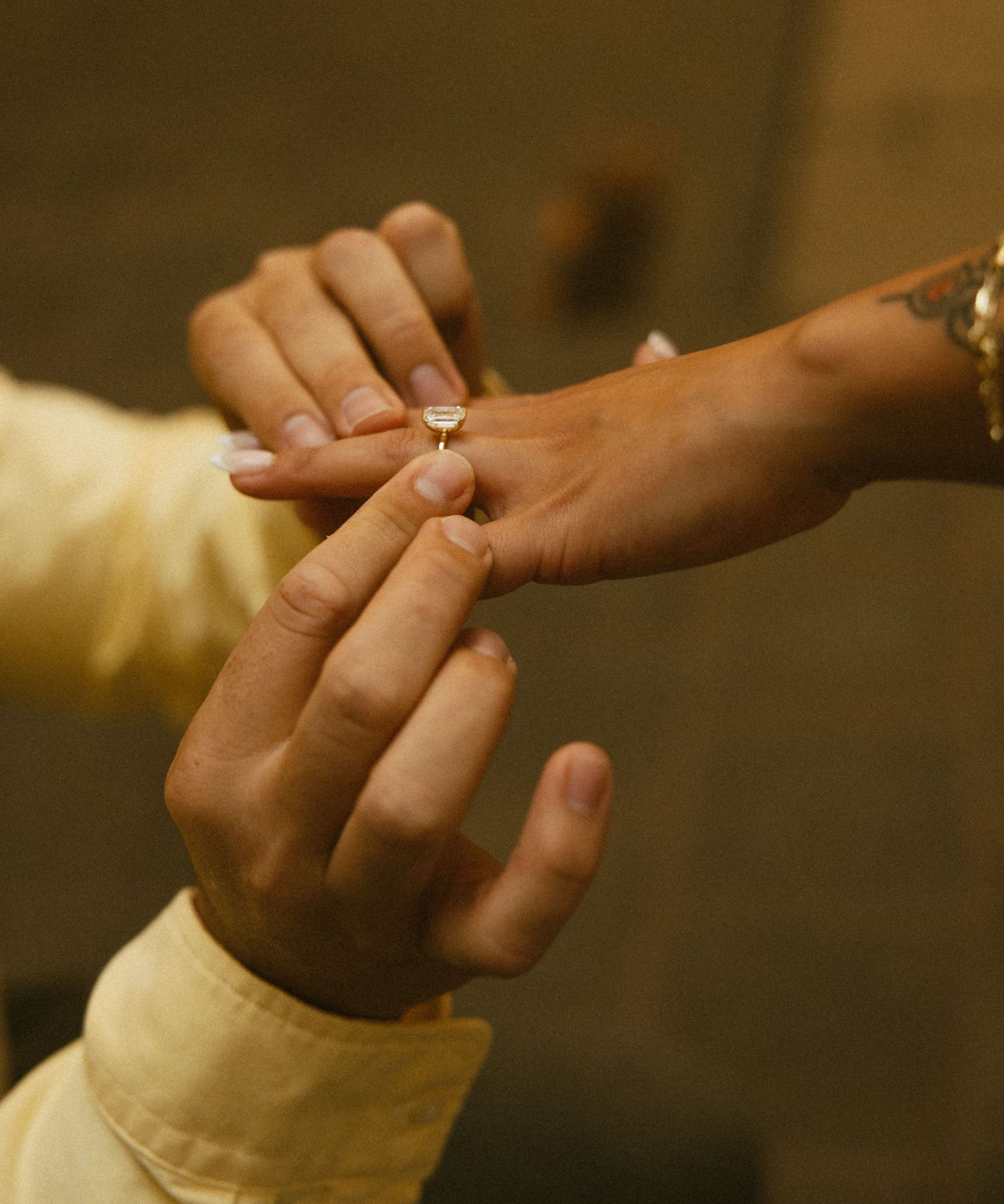 Close-up of a hand sliding a diamond engagement ring onto a woman's finger, her wrist showing a small tattoo.