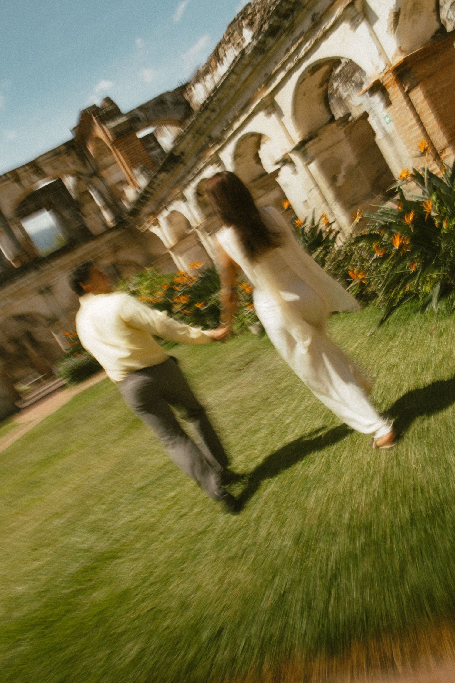 Couple holding hands and walking across a sunny green lawn toward historic stone ruins with arches and orange flowers.