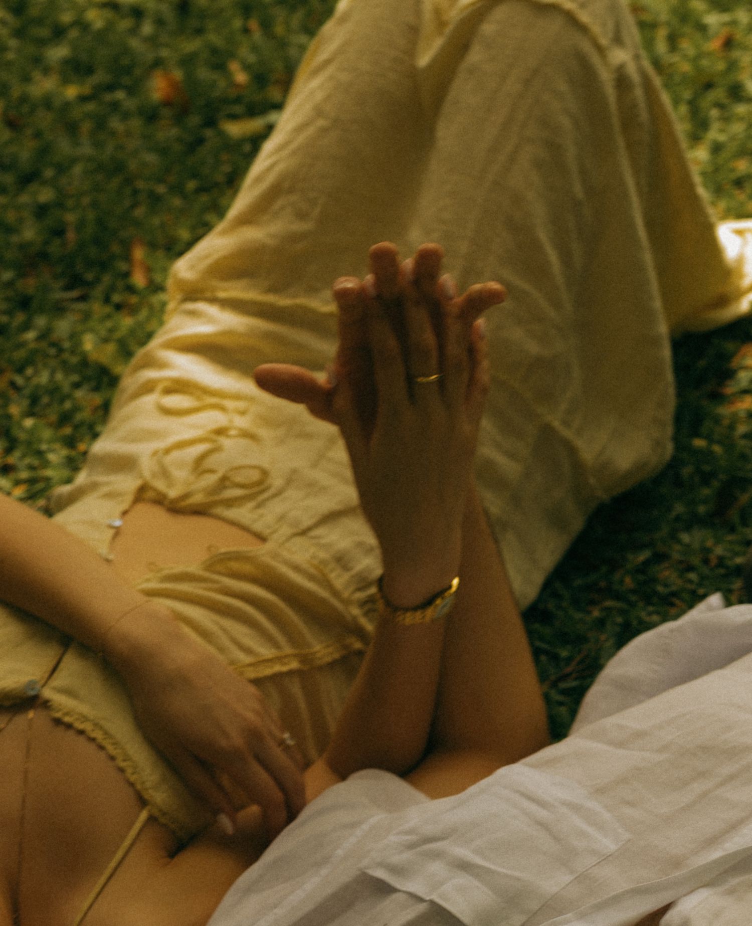 Two people lying on grass, hands intertwined, wearing yellow linen clothing with gold bracelet and ring.