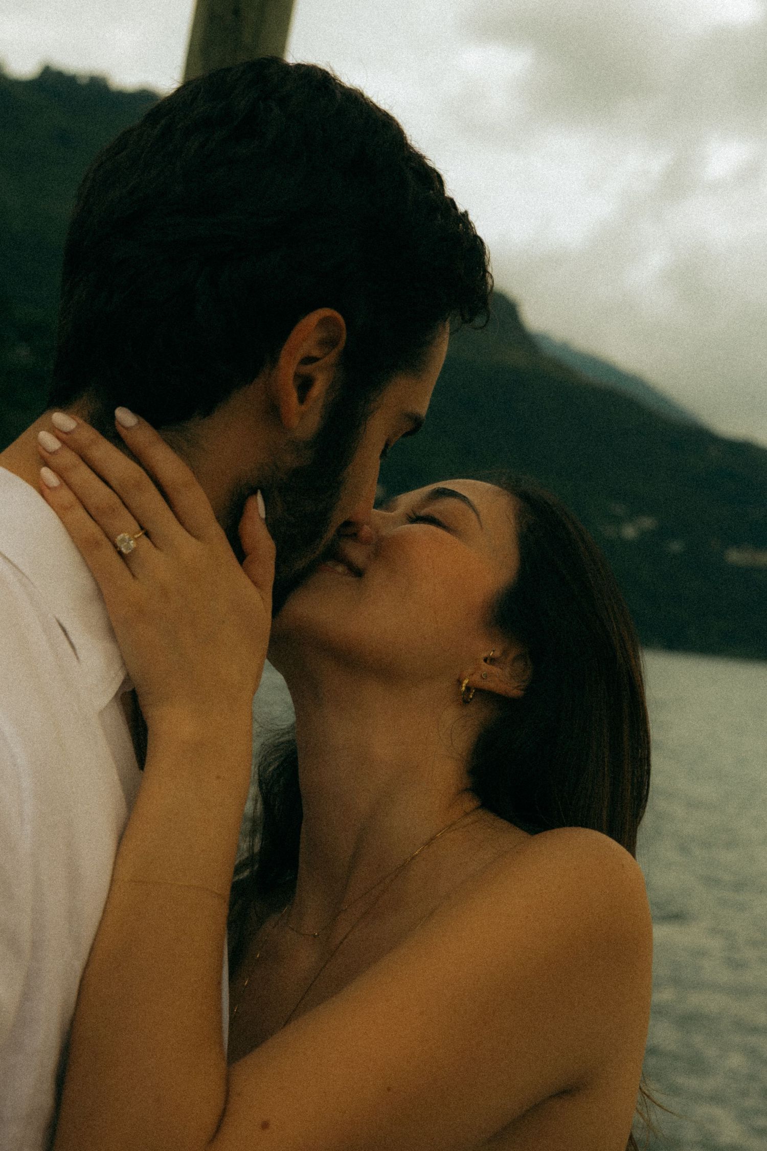 Romantic couple sharing a tender kiss with scenic mountain lake backdrop in moody outdoor engagement photo.