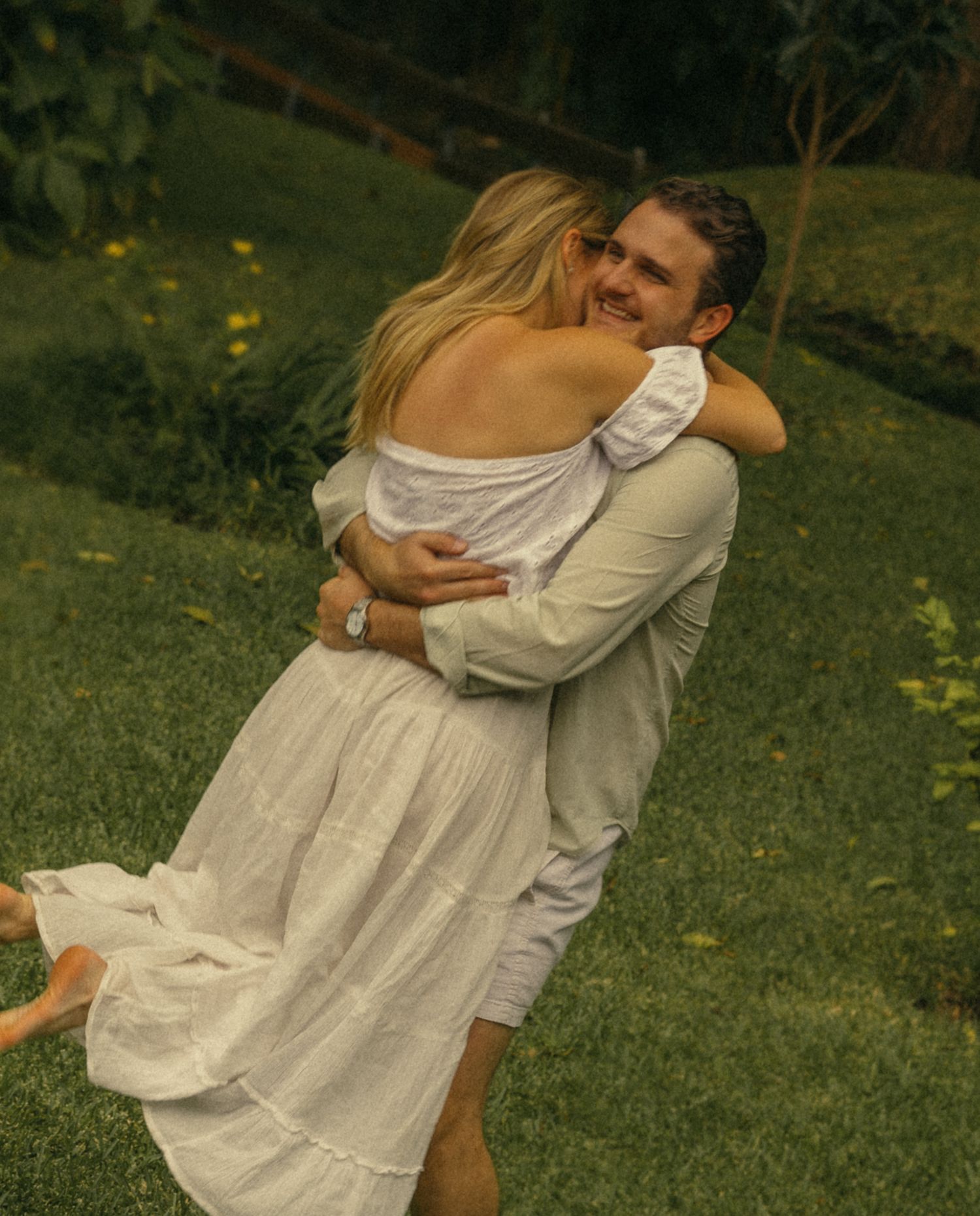 Couple in white outfits embracing and smiling on a lush green lawn in a romantic outdoor setting.